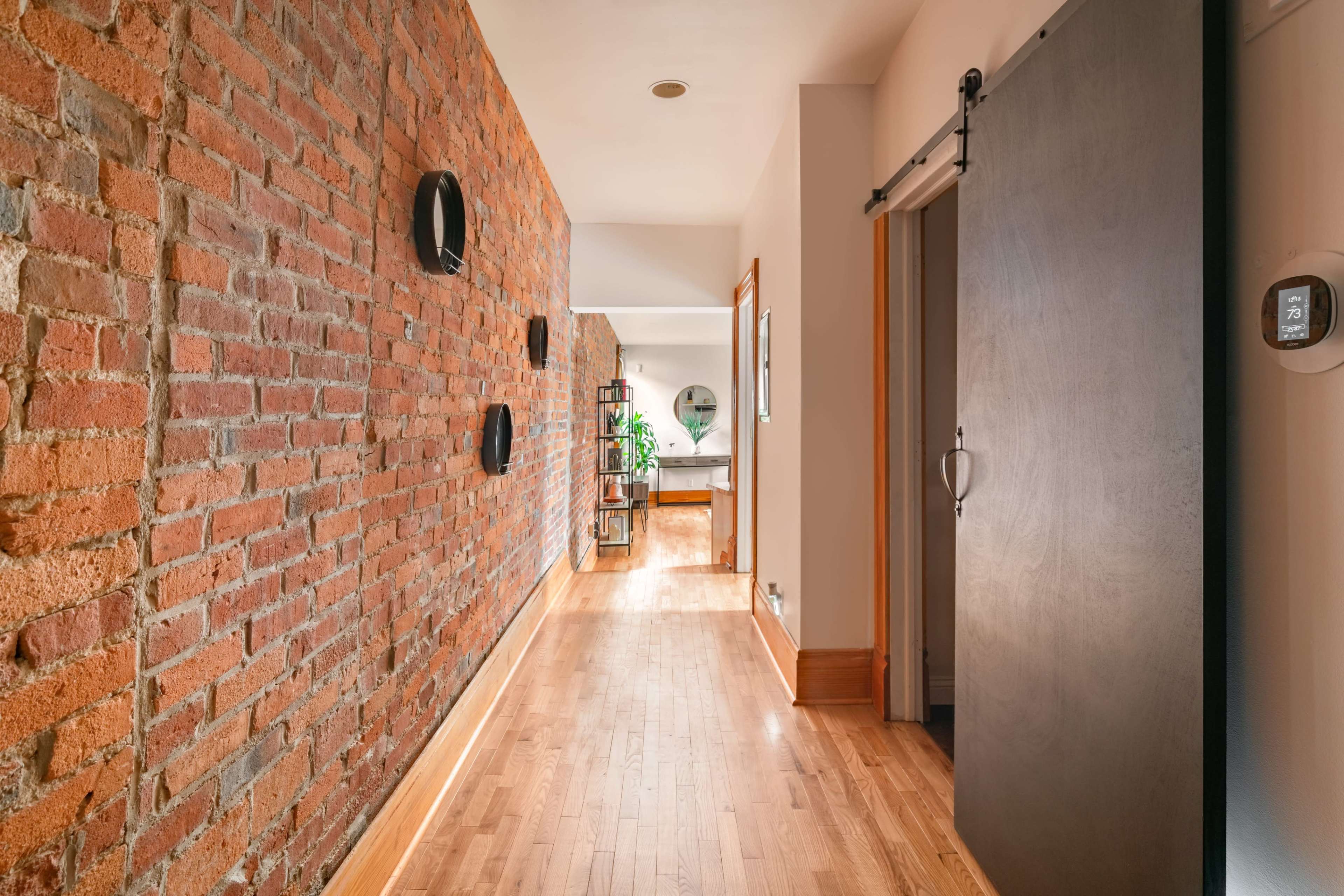 A corridor with a brick wall on one side, wooden flooring, and a large sliding dark door leading to a room.