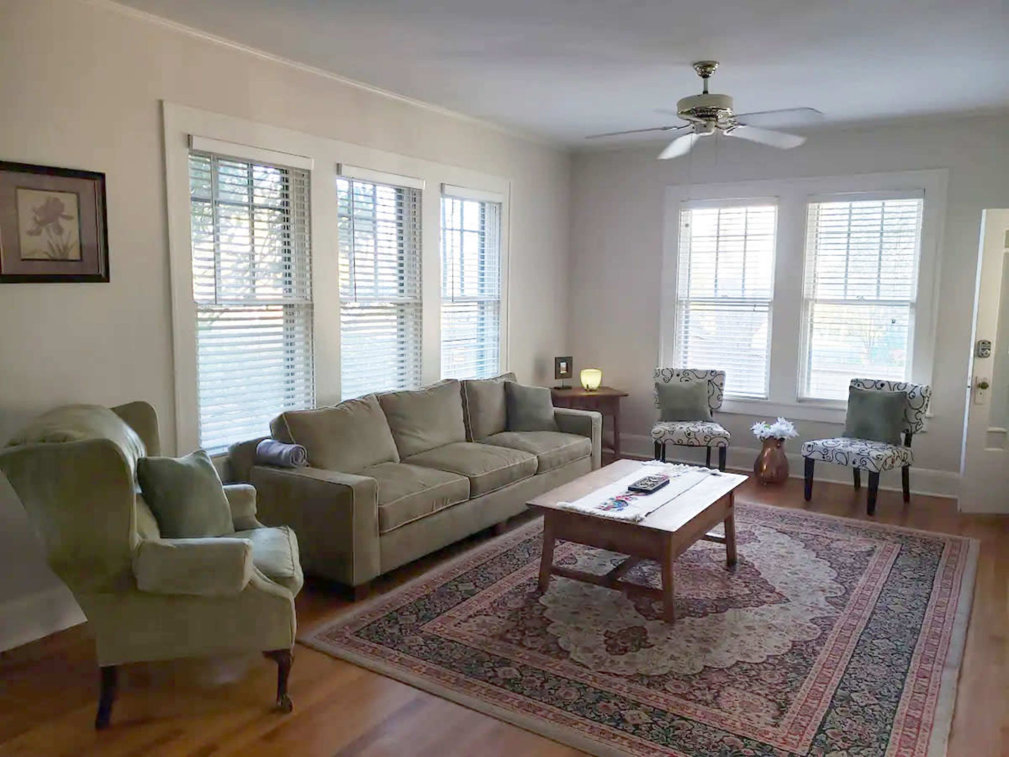 The room features a green sofa, two patterned chairs, a wooden coffee table, and large windows with blinds.