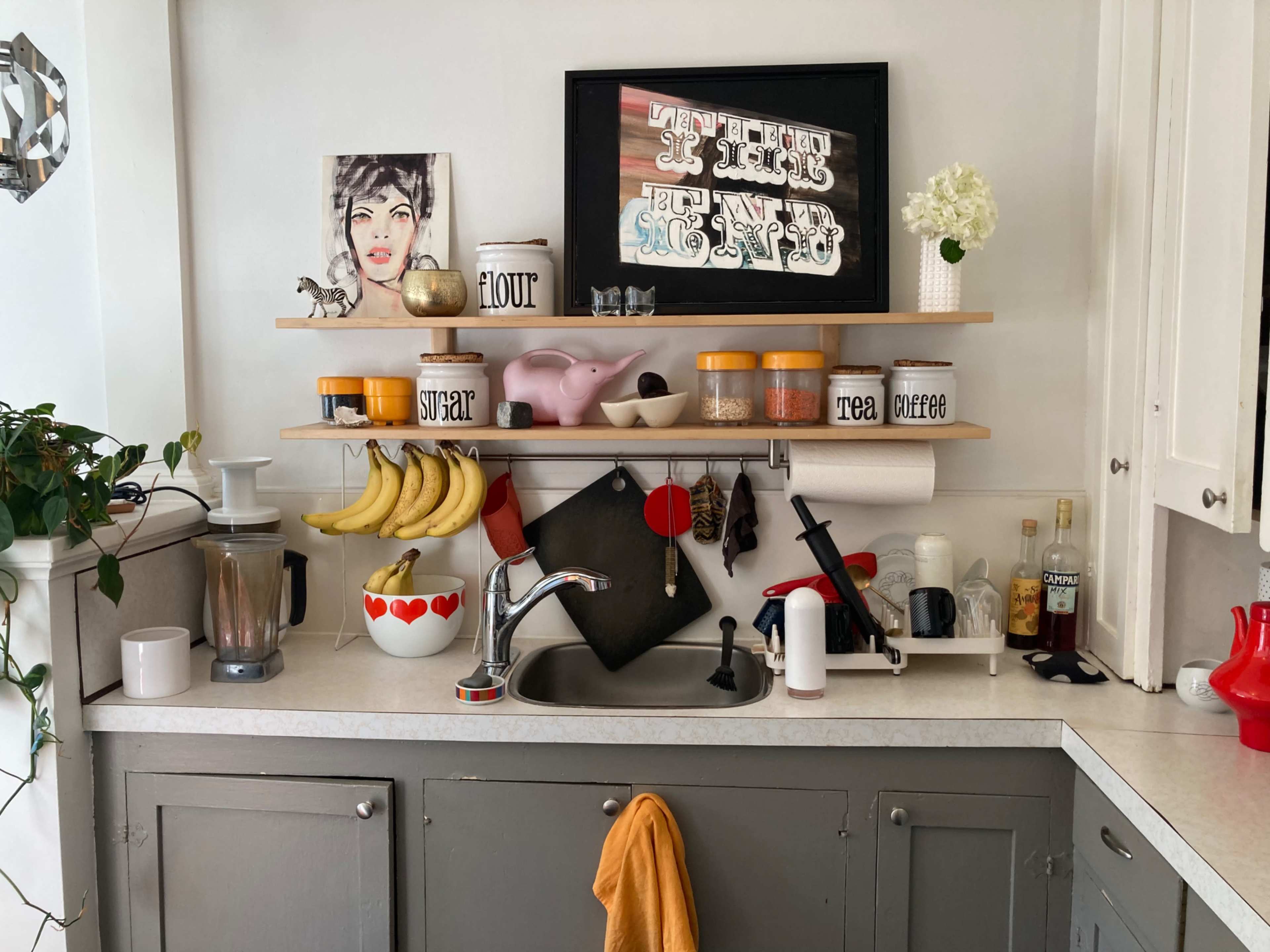 A kitchen counter features a blender, a dish rack, jars labeled with ingredients, a shelf with decorative items, and fresh bananas.