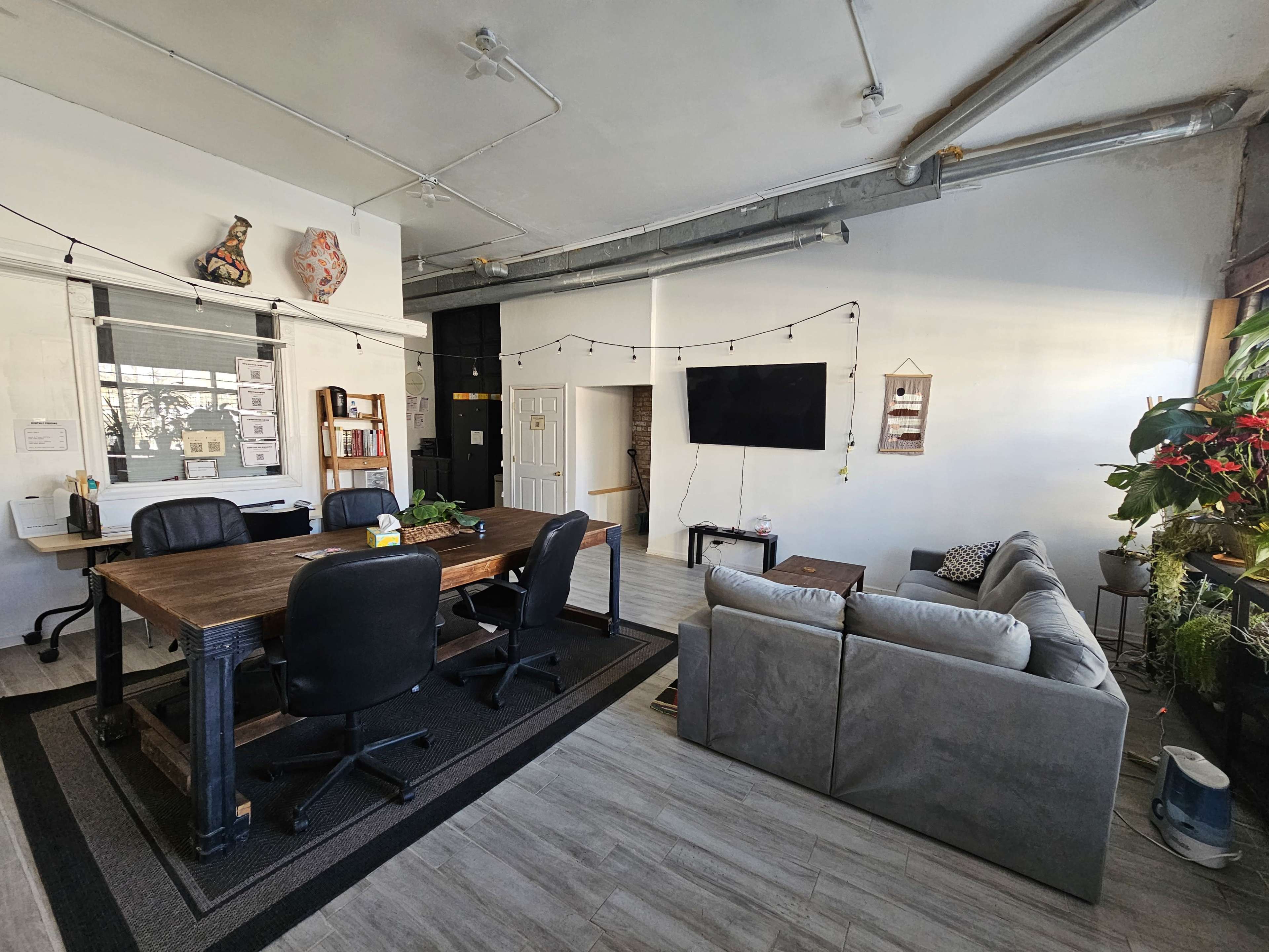 The image shows a modern office space with a large wooden conference table surrounded by black chairs, a gray sofa, and various plants along the perimeter.