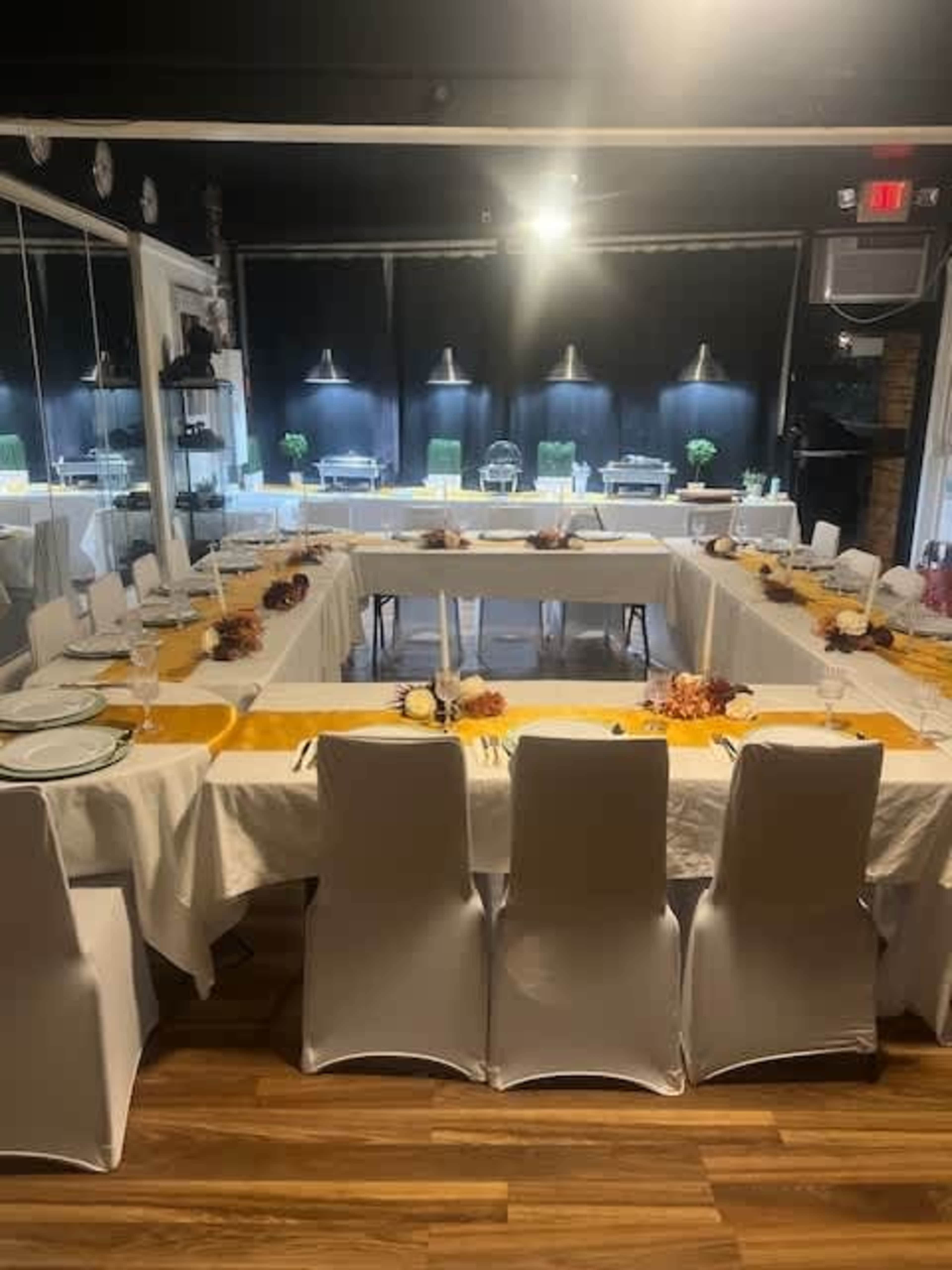A long table is set for a gathering, adorned with white linens and decorative elements, surrounded by white chairs in a well-lit room.