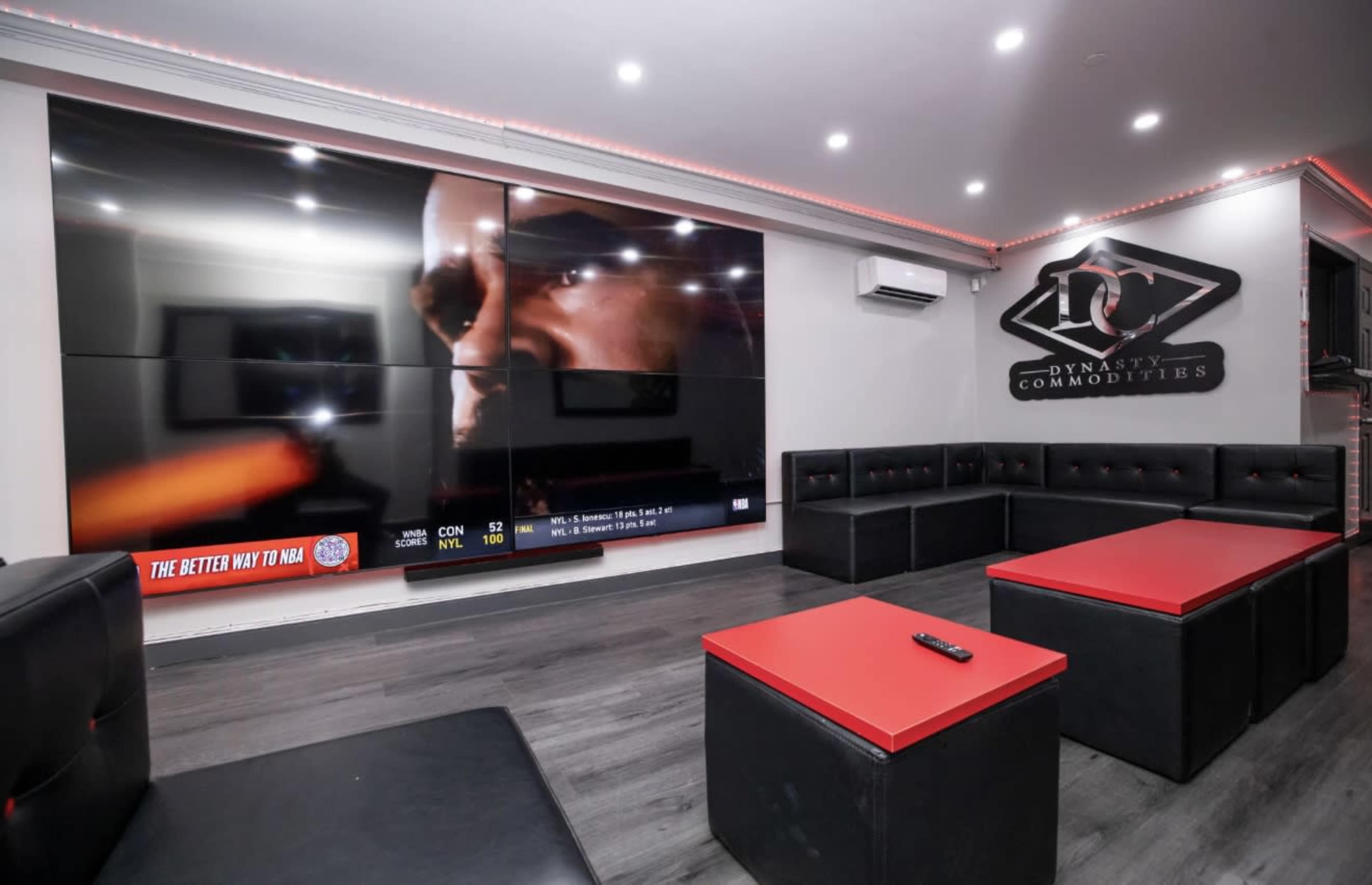 The image shows a modern entertainment room with large screens on the wall, black and red seating, and a low table in the center.