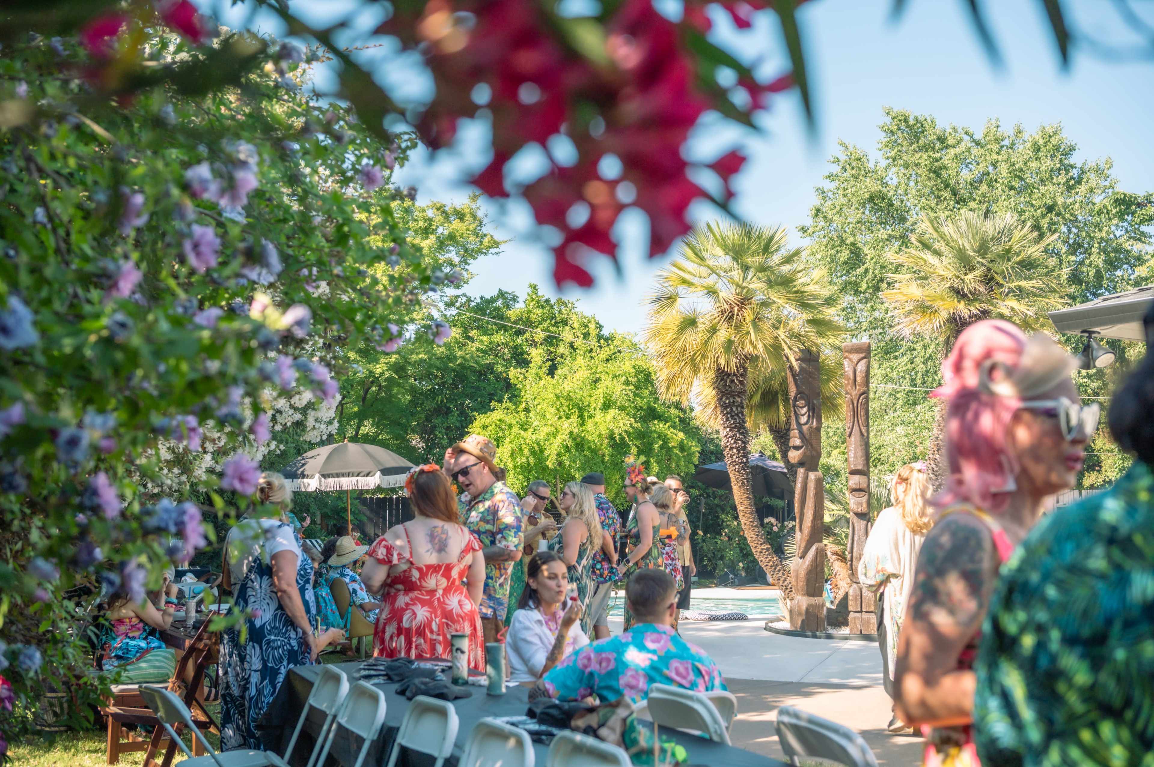 The scene depicts a lively outdoor gathering with people dressed in colorful attire, surrounded by lush greenery and tropical plants.