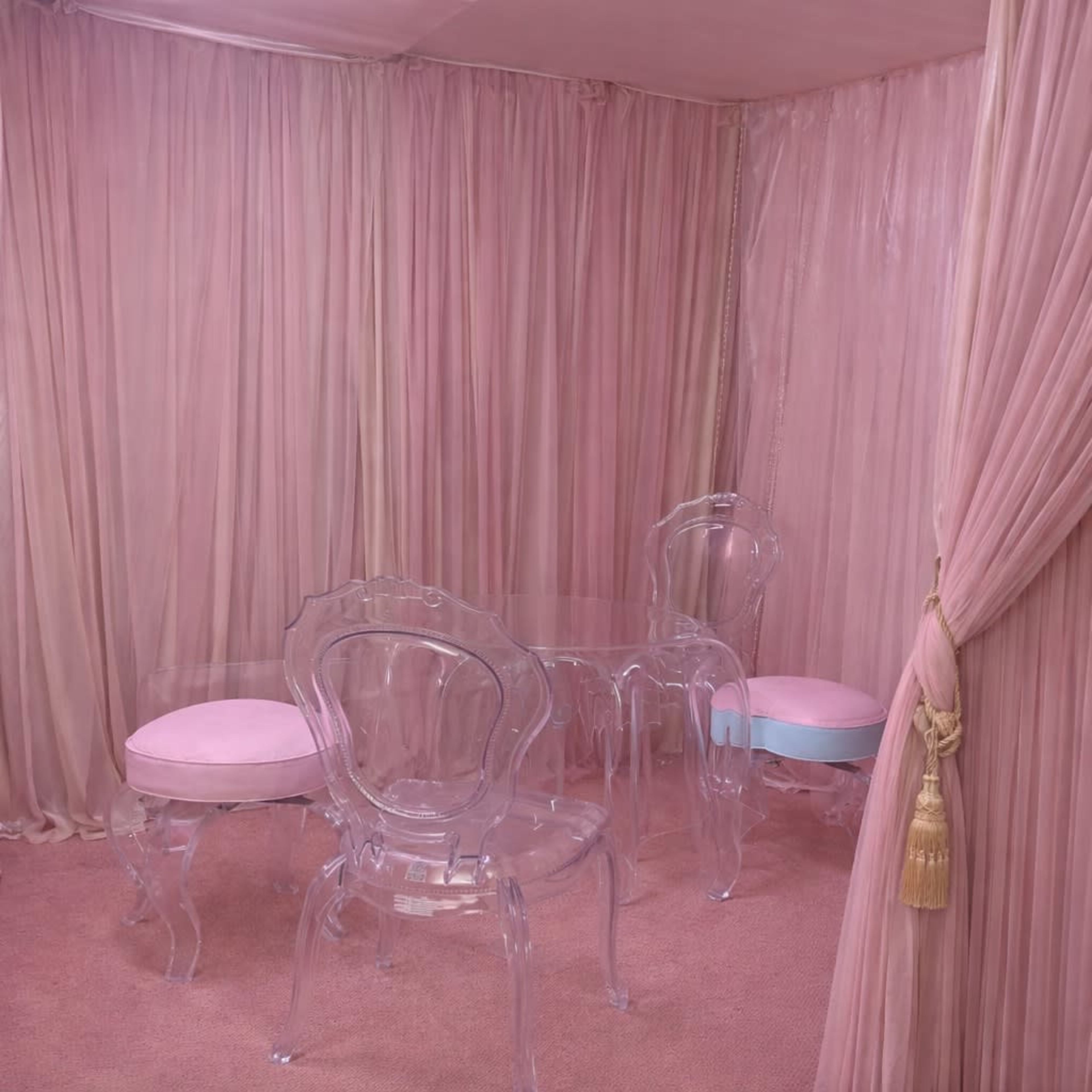 The image shows a room draped in pink fabric with a clear acrylic table and chairs, featuring pink cushions.
