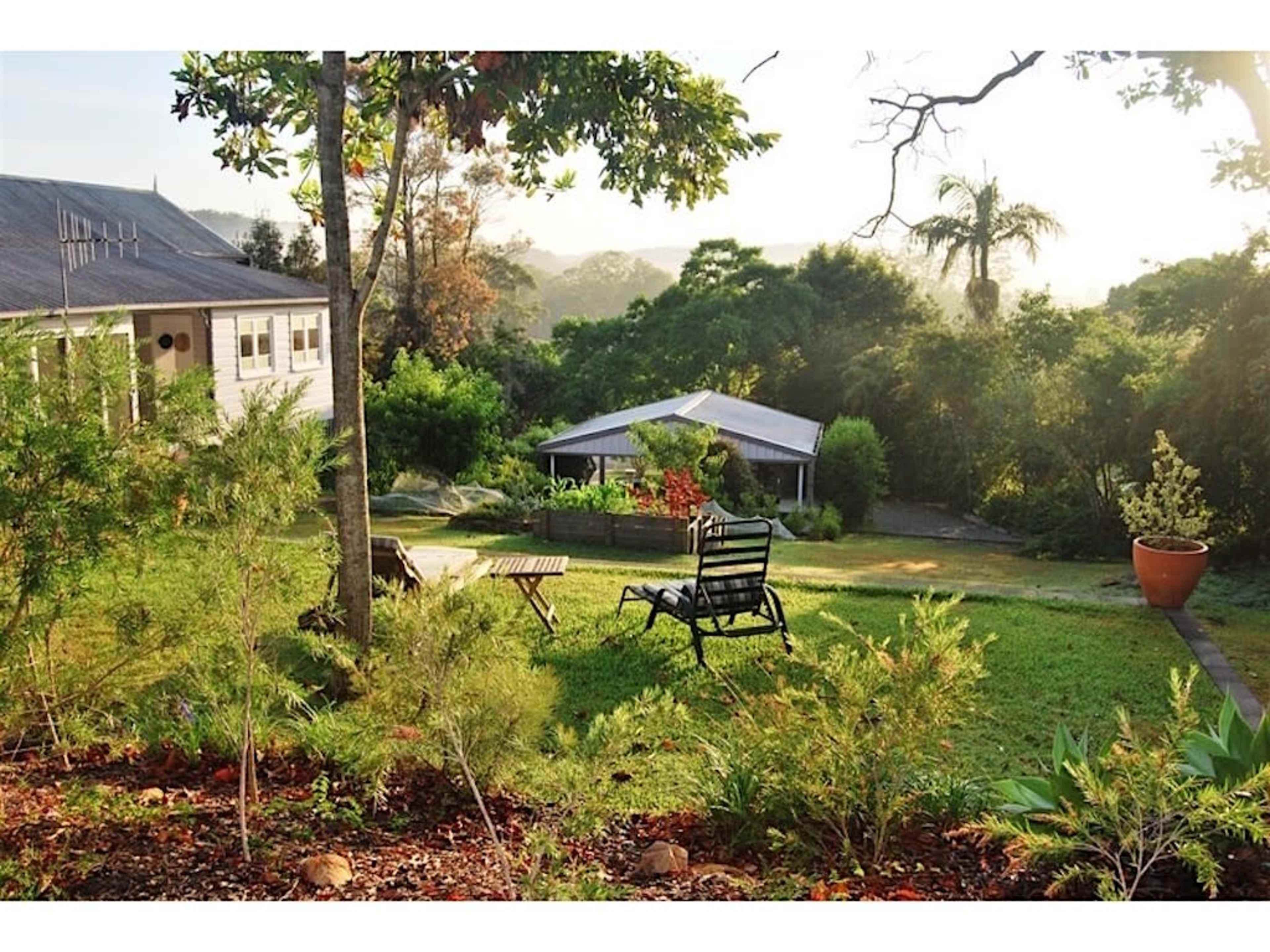 The image shows a garden with a green lawn, a wooden chair, small tables, and a house in the background surrounded by trees and shrubs.