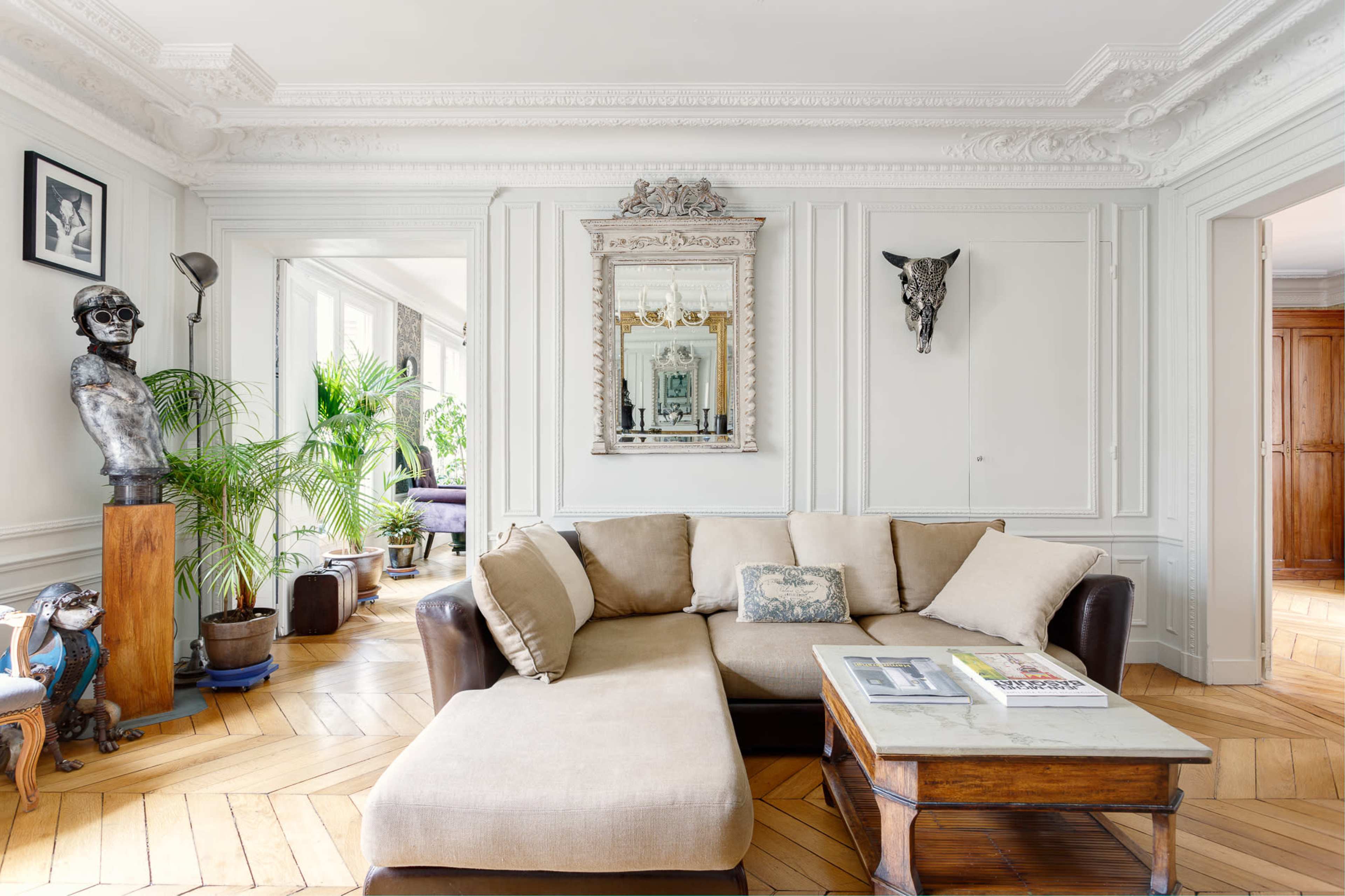 A spacious living room with a beige sectional sofa, a wooden coffee table, and decorative plants, surrounded by white-framed walls and intricate moldings.