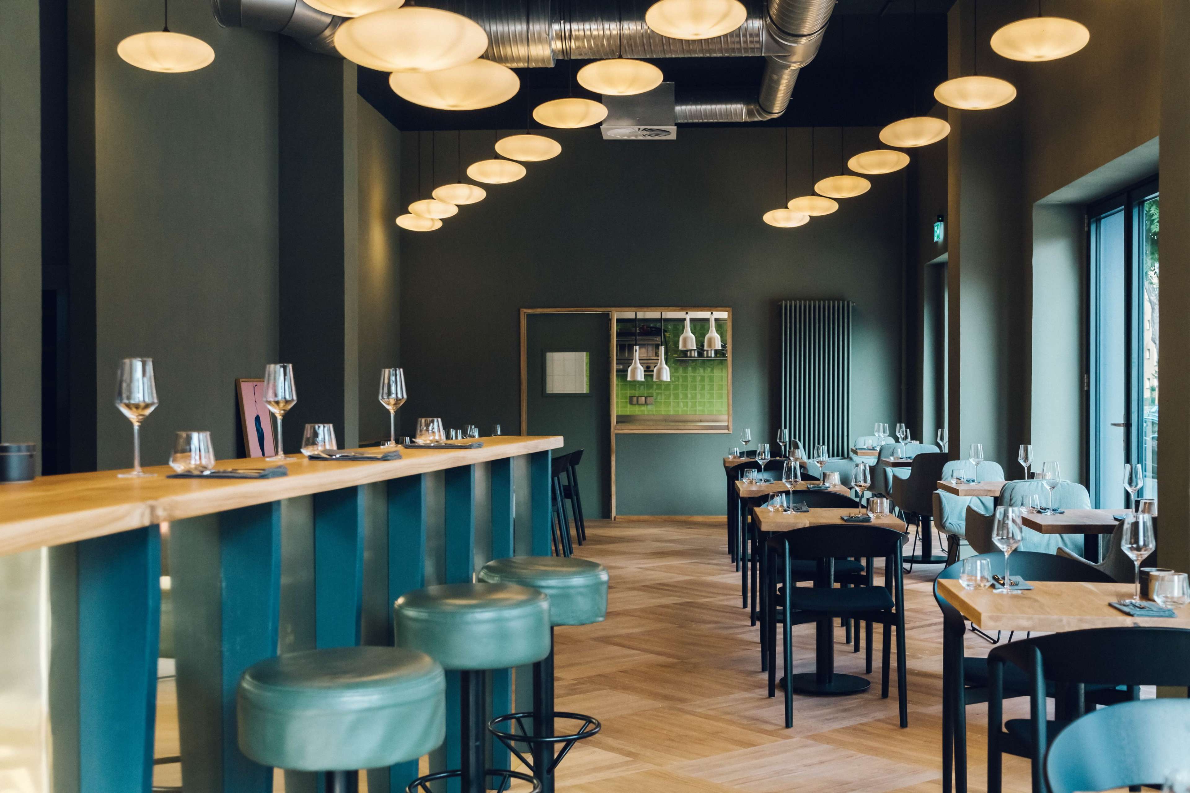 The image shows a modern restaurant interior with green walls, wooden flooring, and several round pendant lights hanging from the ceiling, along with spaced-out tables and bar seating.