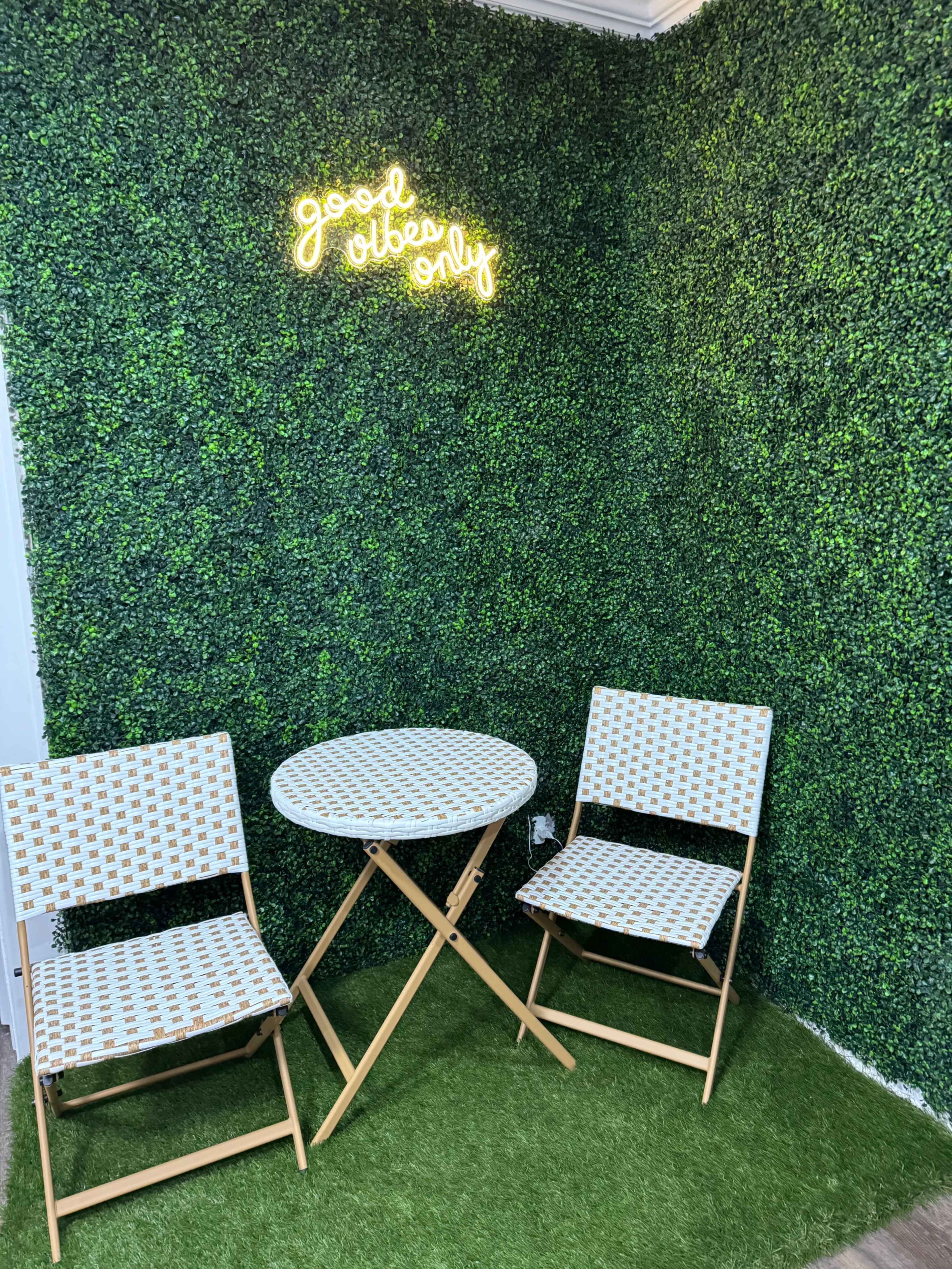 A neon sign reading "good vibes only" illuminates a small seating area with a round table and two patterned chairs against a green wall covered in artificial ivy.
