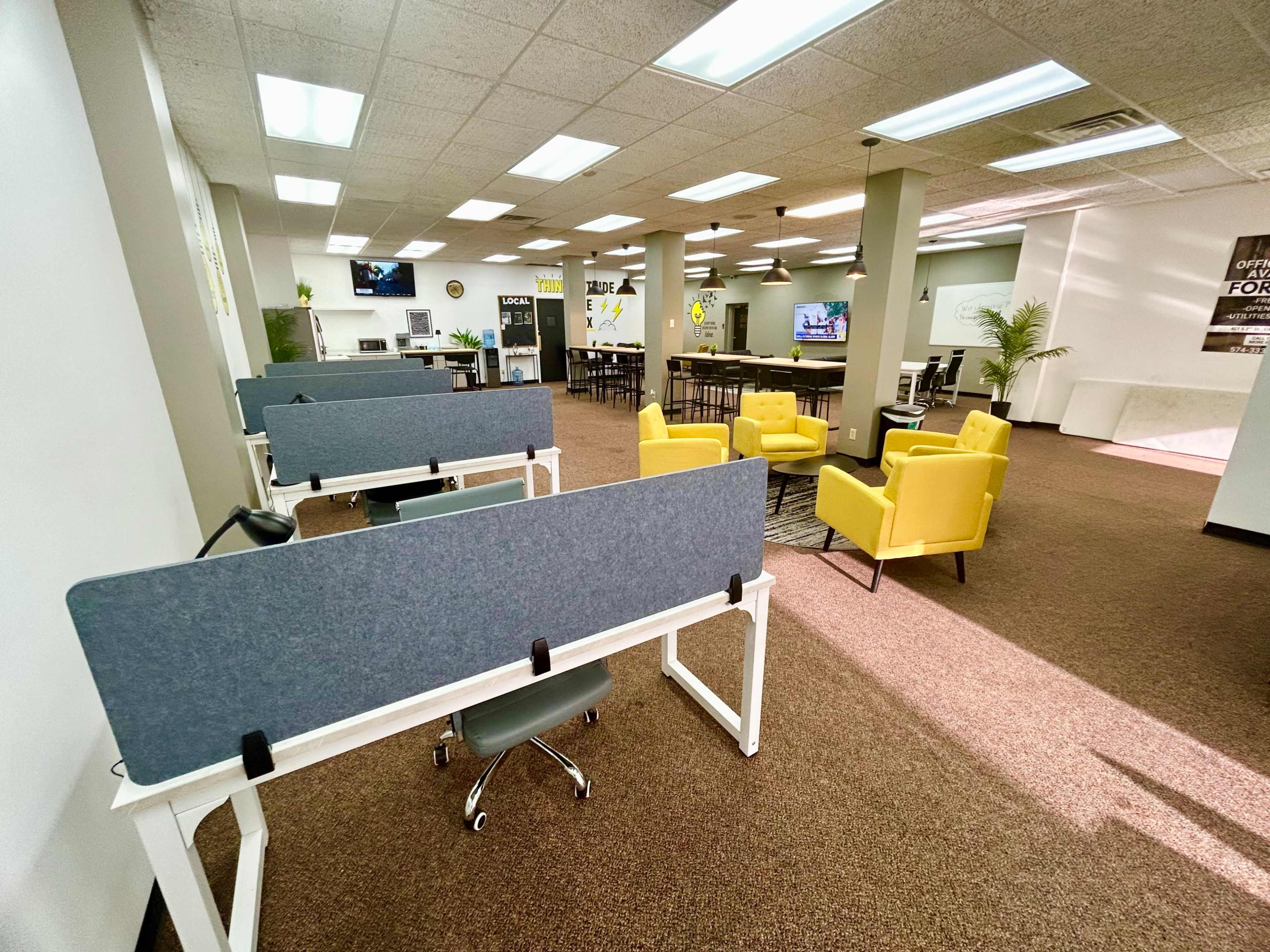 The image shows a spacious office area featuring several workstations with gray dividers, yellow chairs, and a communal seating space with tables and multiple screens.