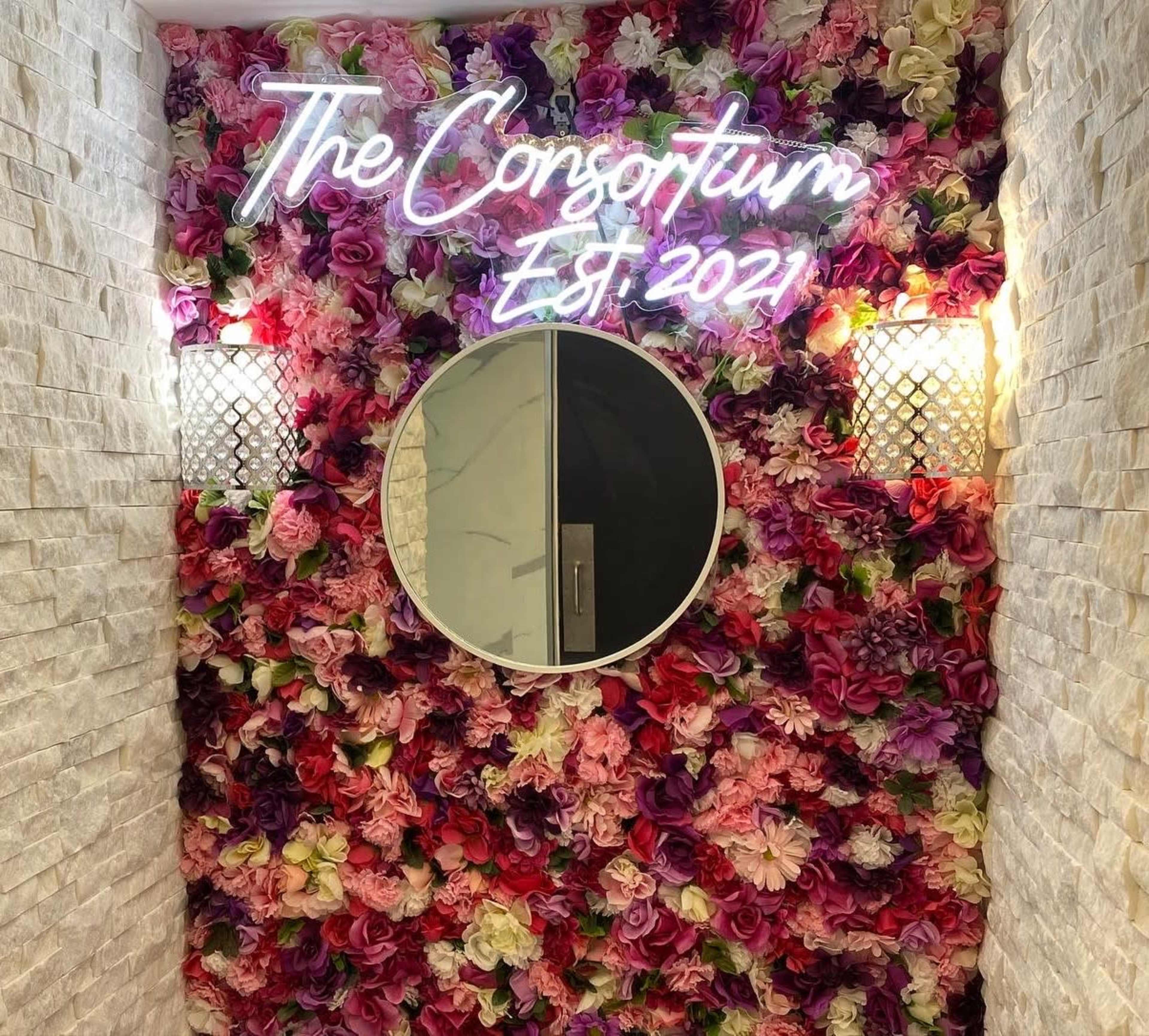 A wall covered in artificial flowers with a round mirror and a neon sign that reads "The Consortium Est. 2021."