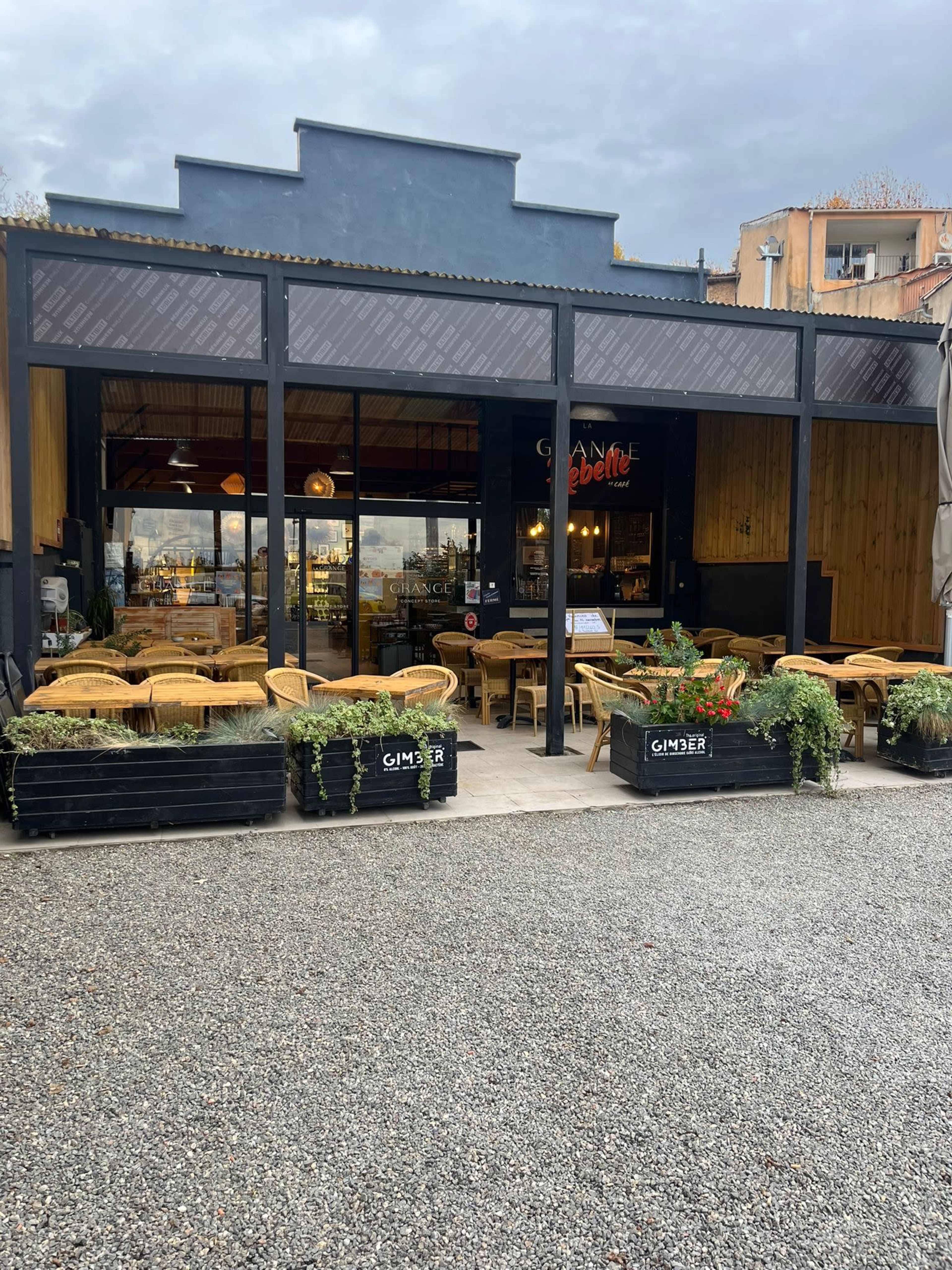 The image shows a modern restaurant with large glass doors and an outdoor seating area featuring wicker chairs and potted plants.