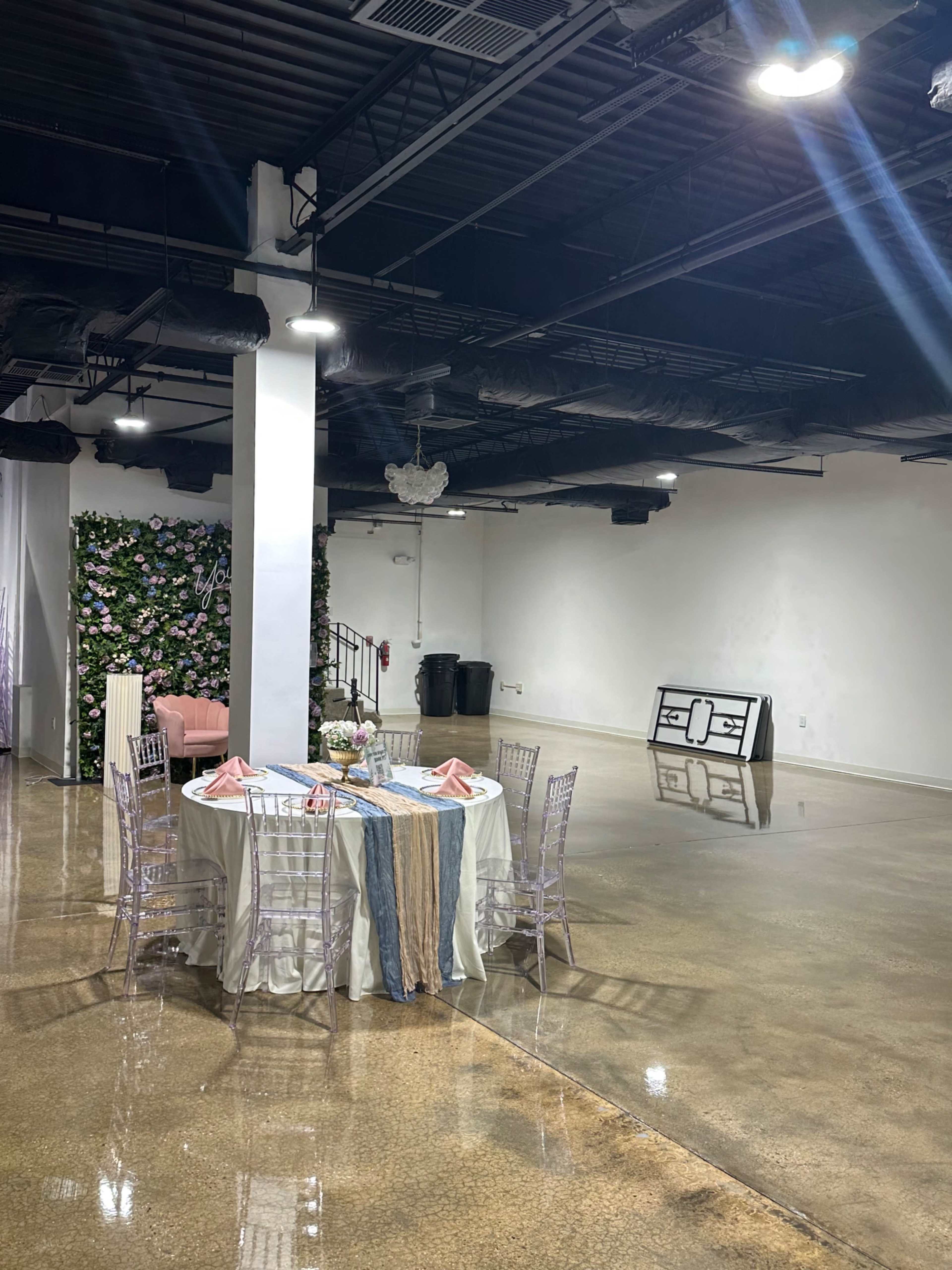 A large, empty event space features a round dining table set with decorative linens, surrounded by clear chairs, and a floral backdrop against one wall.