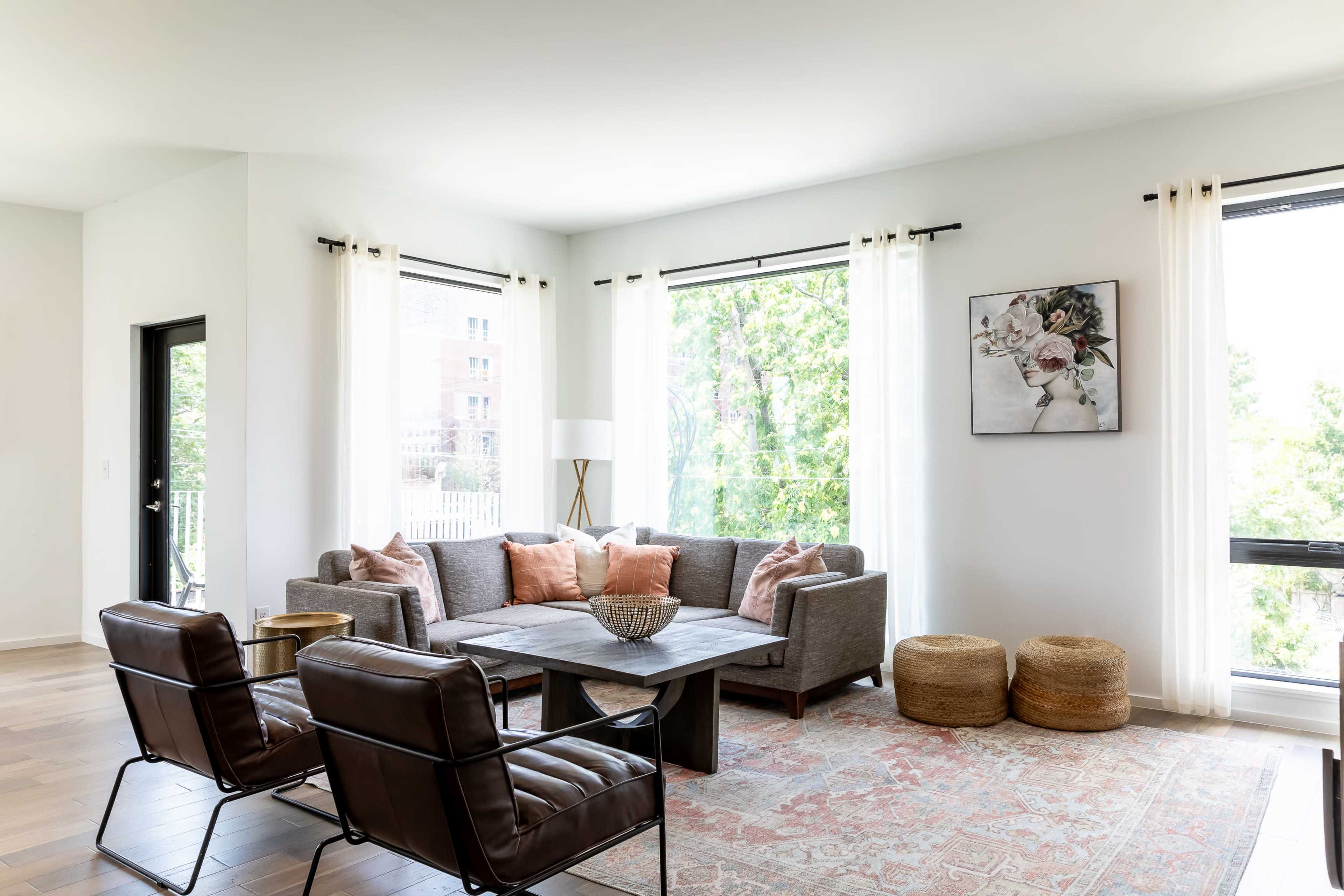 A modern living room features a gray sectional sofa, two leather chairs, a wooden coffee table, and large windows with sheer curtains.