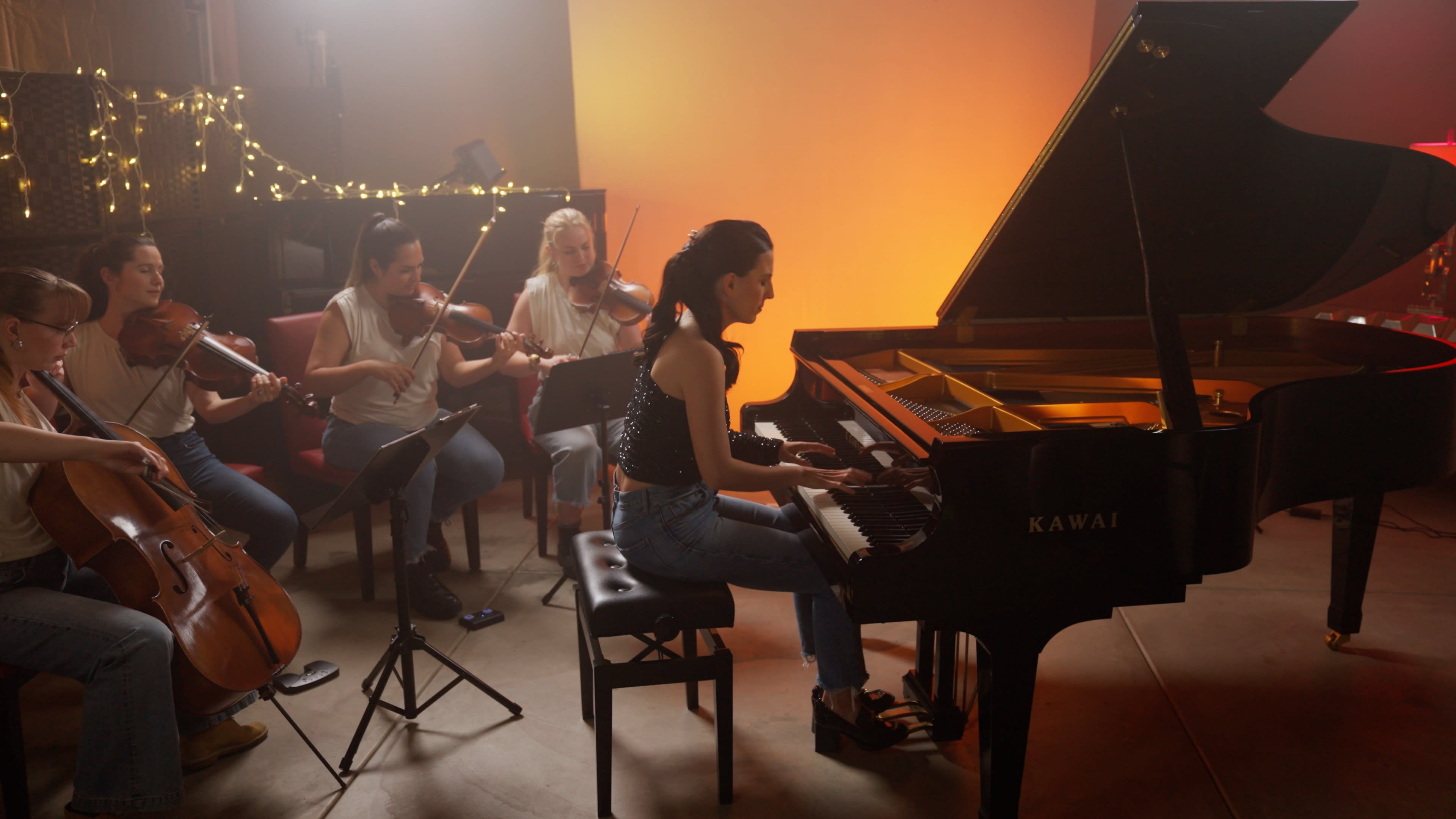 A pianist plays at a grand piano while four musicians, including violinists and a cellist, perform in a well-lit studio.