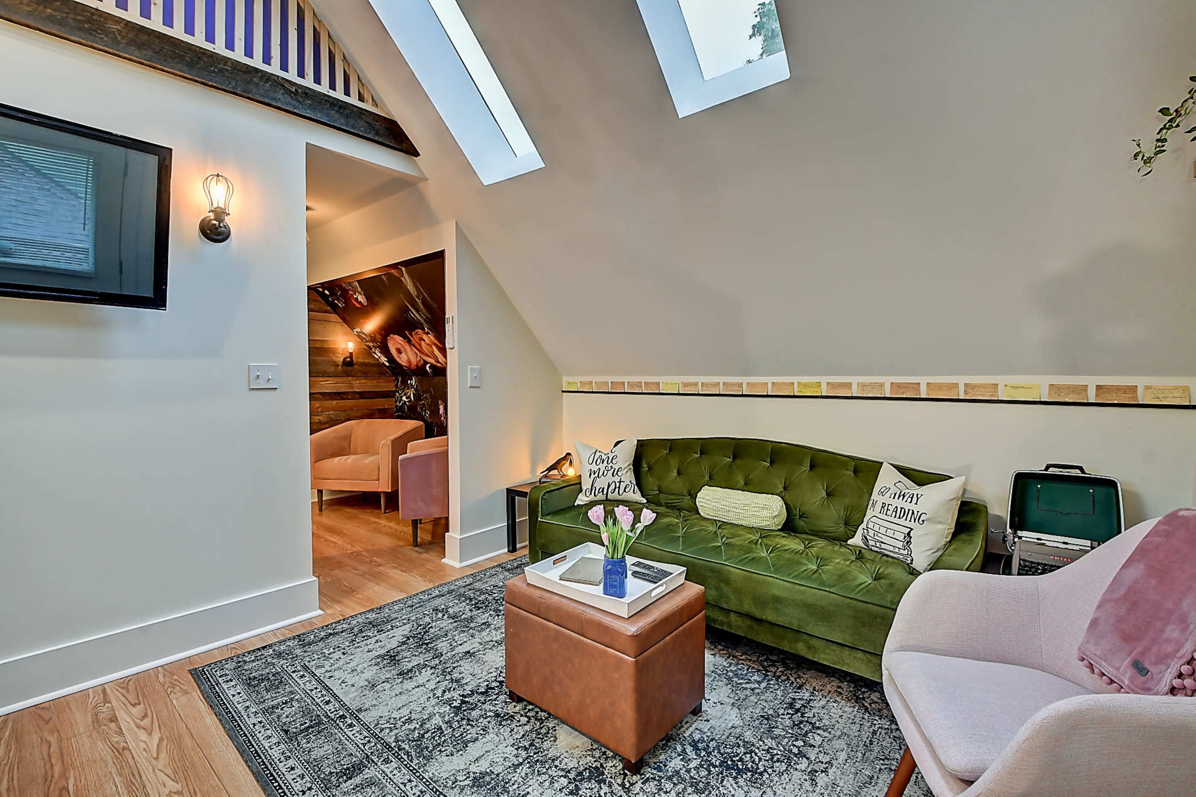 The image shows a cozy living space featuring a green sofa, a coffee table, and a pink chair, with skylights and a decorative wall.