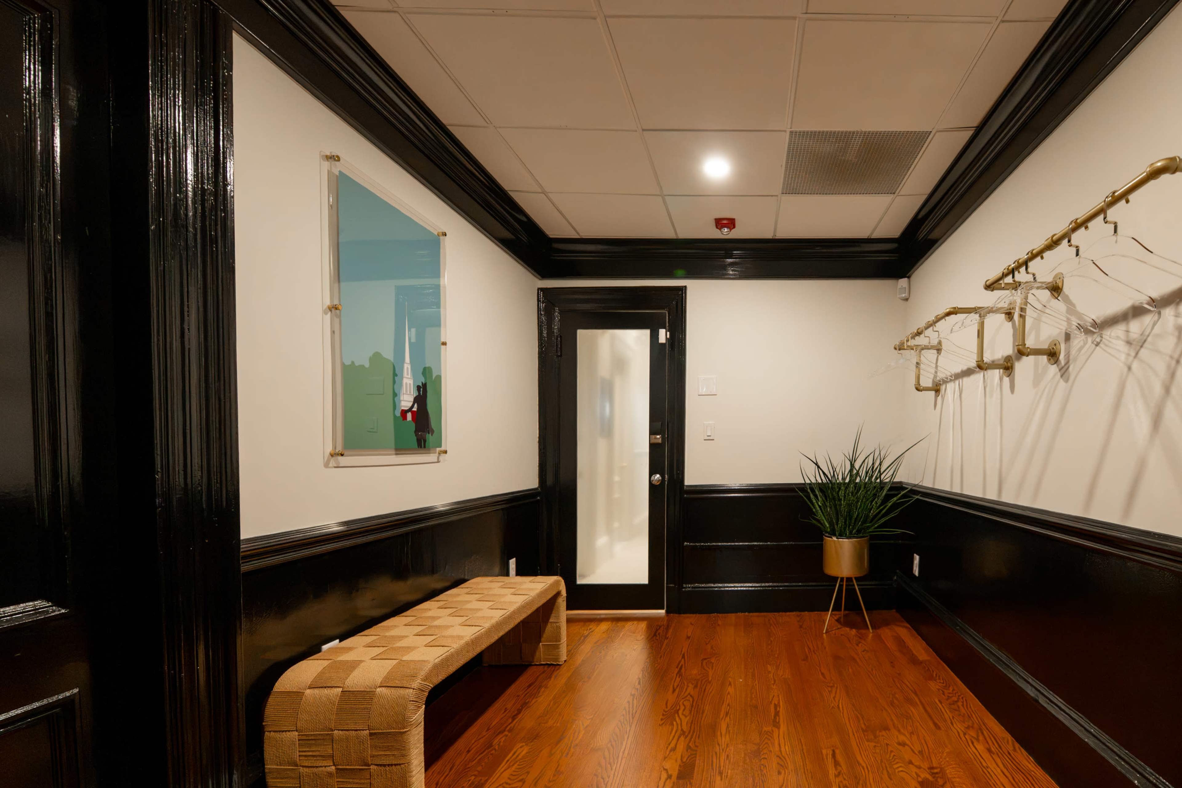 The image shows a hallway with a woven bench, a potted plant, a door with a glass panel, and wall hooks against a backdrop of dark wood and white walls.