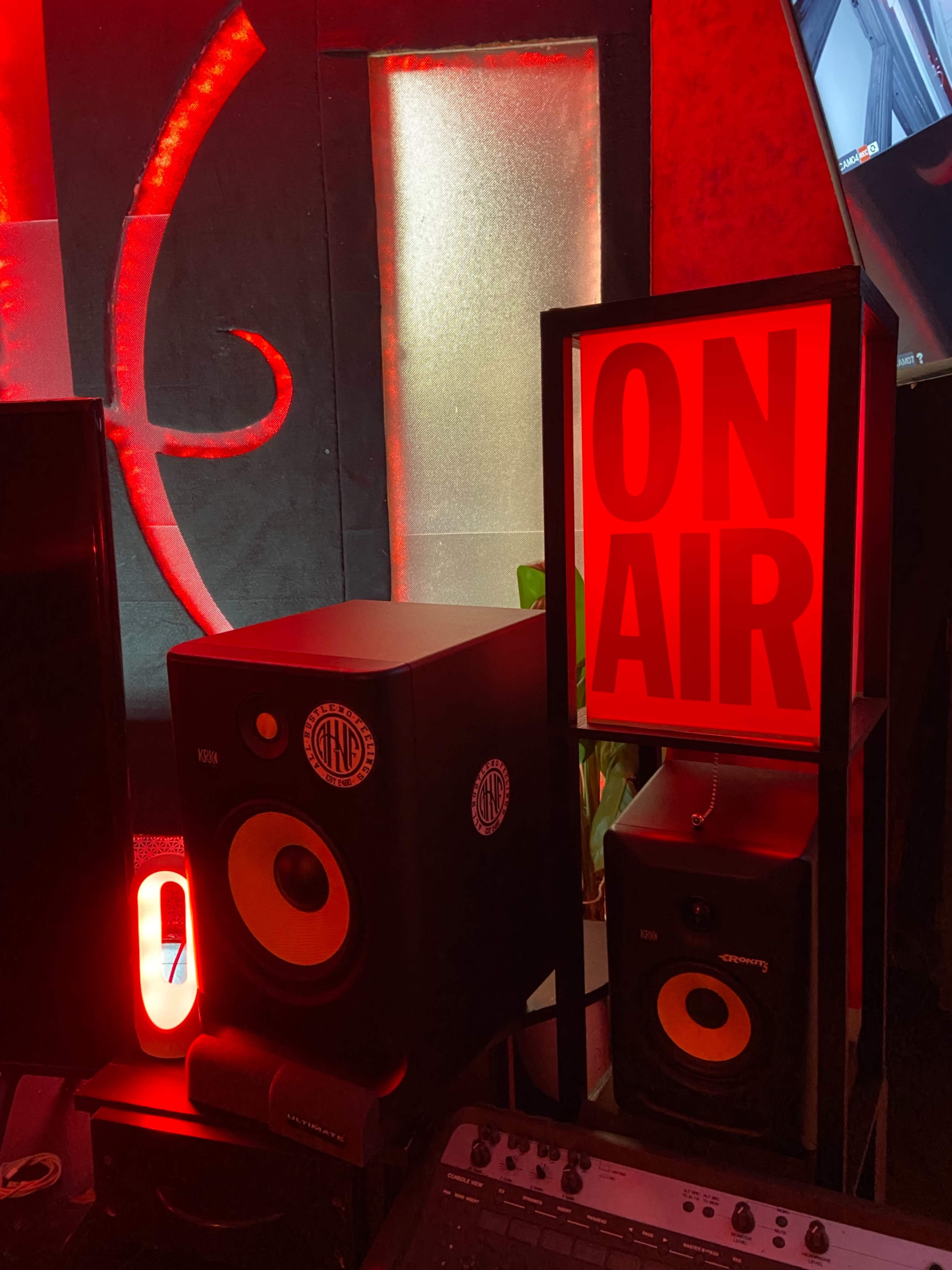 The image shows a recording studio setup with black speakers, a glowing "ON AIR" sign, and ambient red lighting.