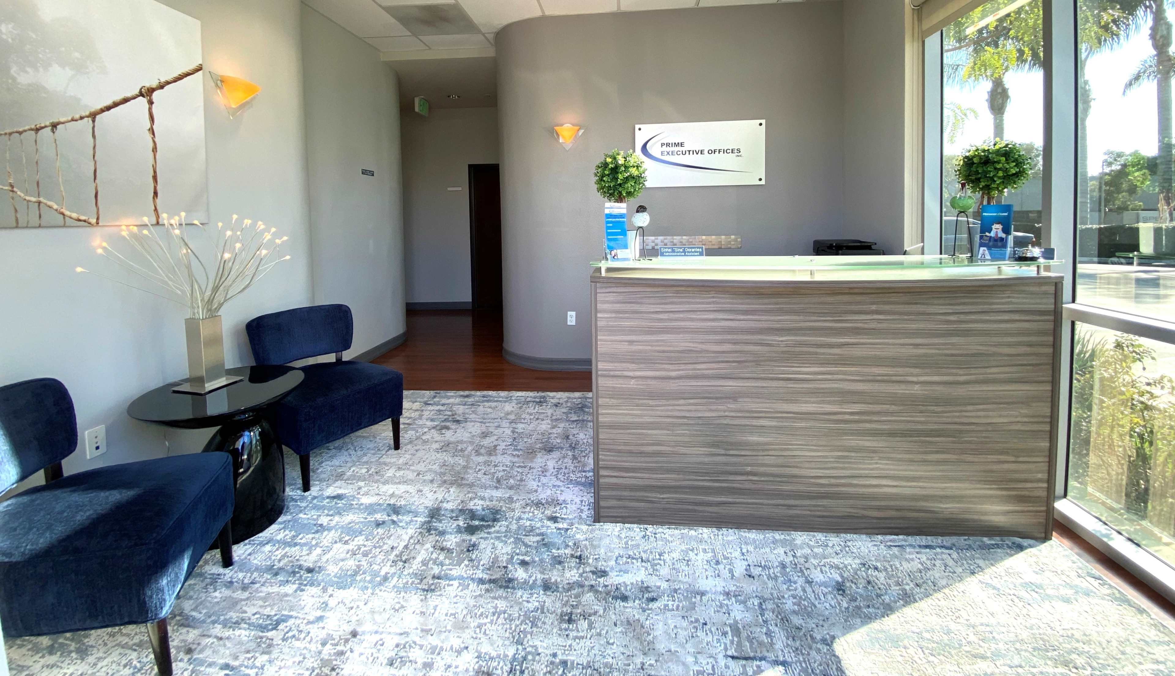 The image shows a reception area with two chairs, a small table, and a front desk, surrounded by light-colored walls and large windows.