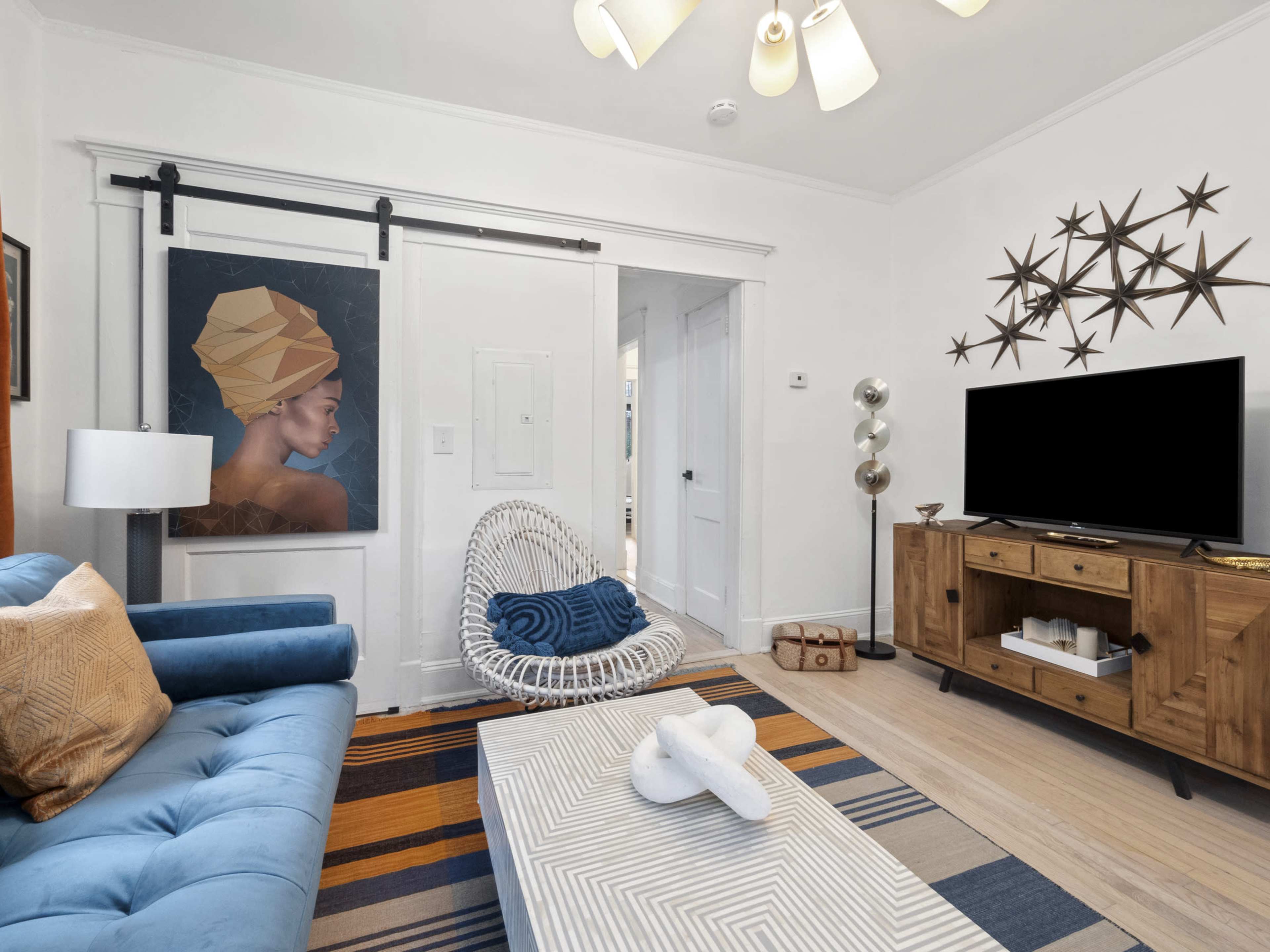 The image shows a living room with a blue sofa, a large framed artwork of a woman on the wall, a white wicker chair, a wooden coffee table, and a television on a wooden stand.