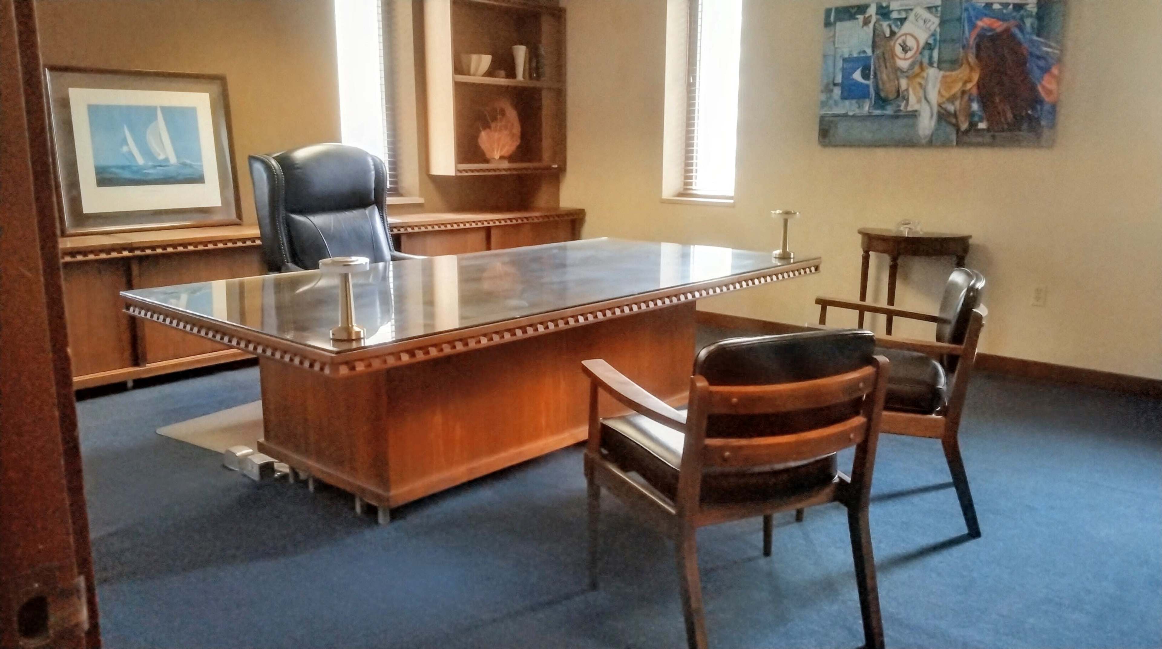The image shows a well-furnished office with a large wooden desk, two chairs, and shelves displaying decorative items and artwork on the walls.