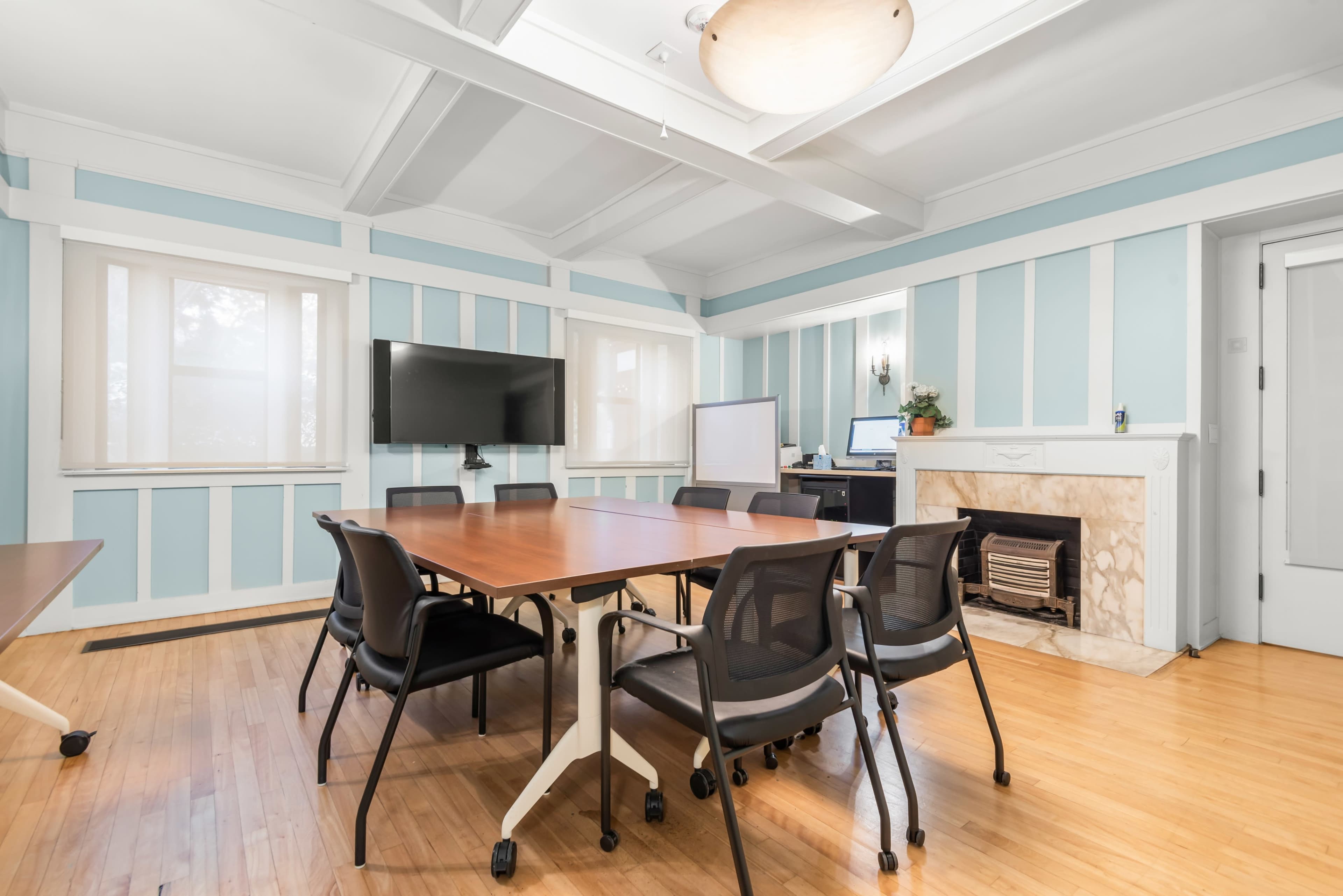 The image shows a meeting room featuring several wooden tables, black rolling chairs, a television screen, a whiteboard, and a fireplace, all set against light blue and white-striped walls.