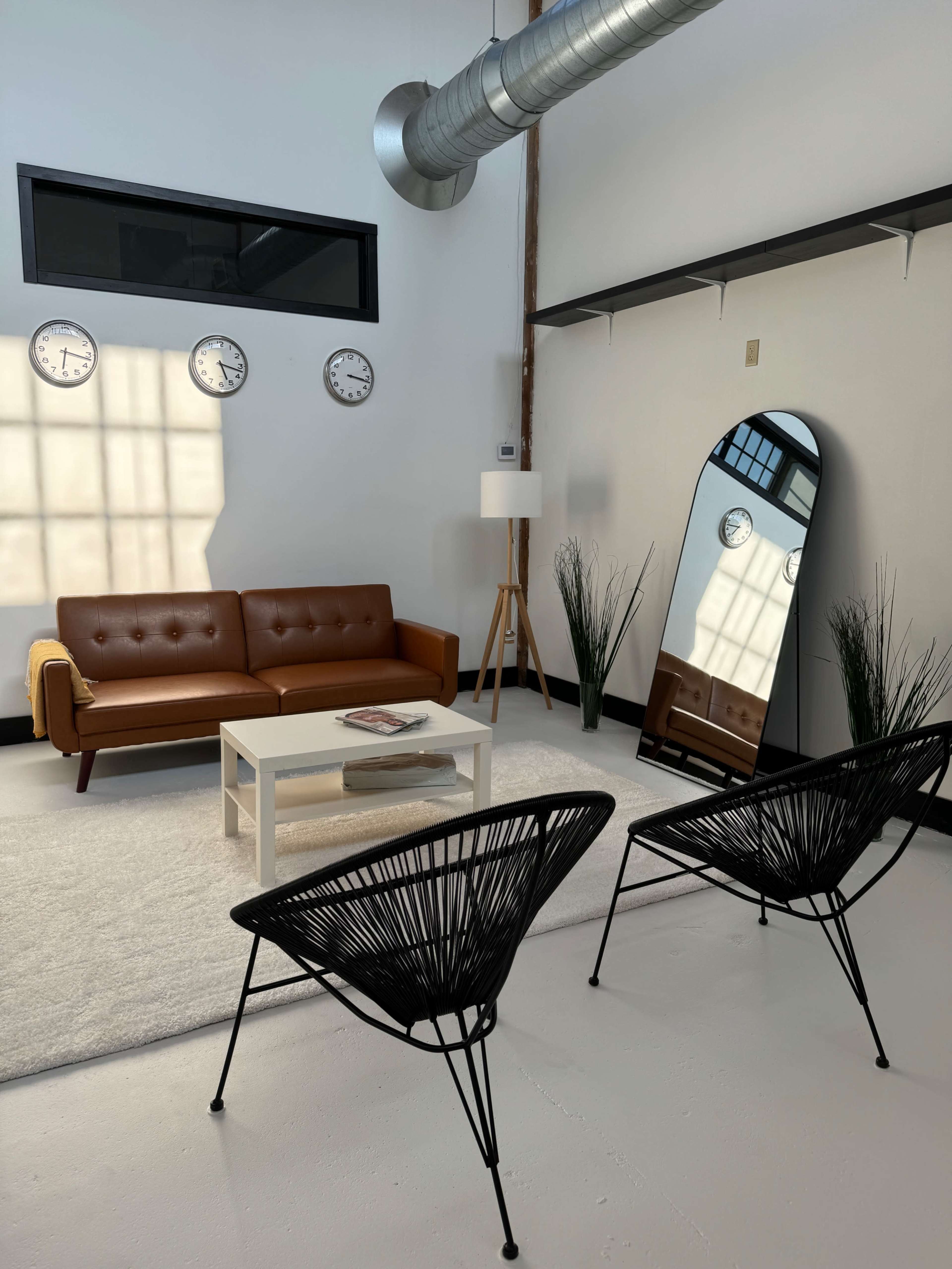 A modern living room featuring a brown leather sofa, a white coffee table, two black chairs, a large mirror, and three wall clocks, all set against a light-colored floor and walls.
