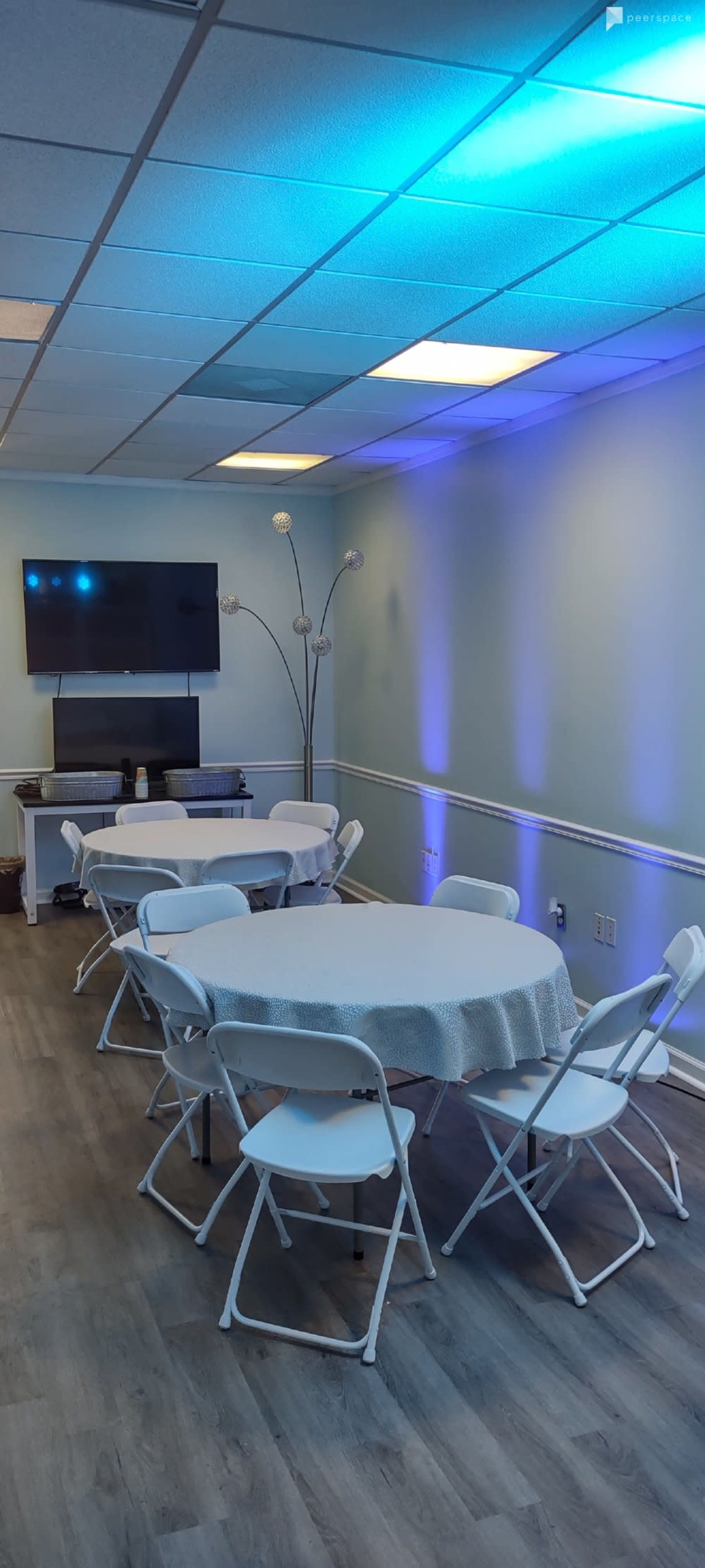 A small meeting room features several round tables surrounded by white chairs, with a television mounted on the wall and accented lighting.
