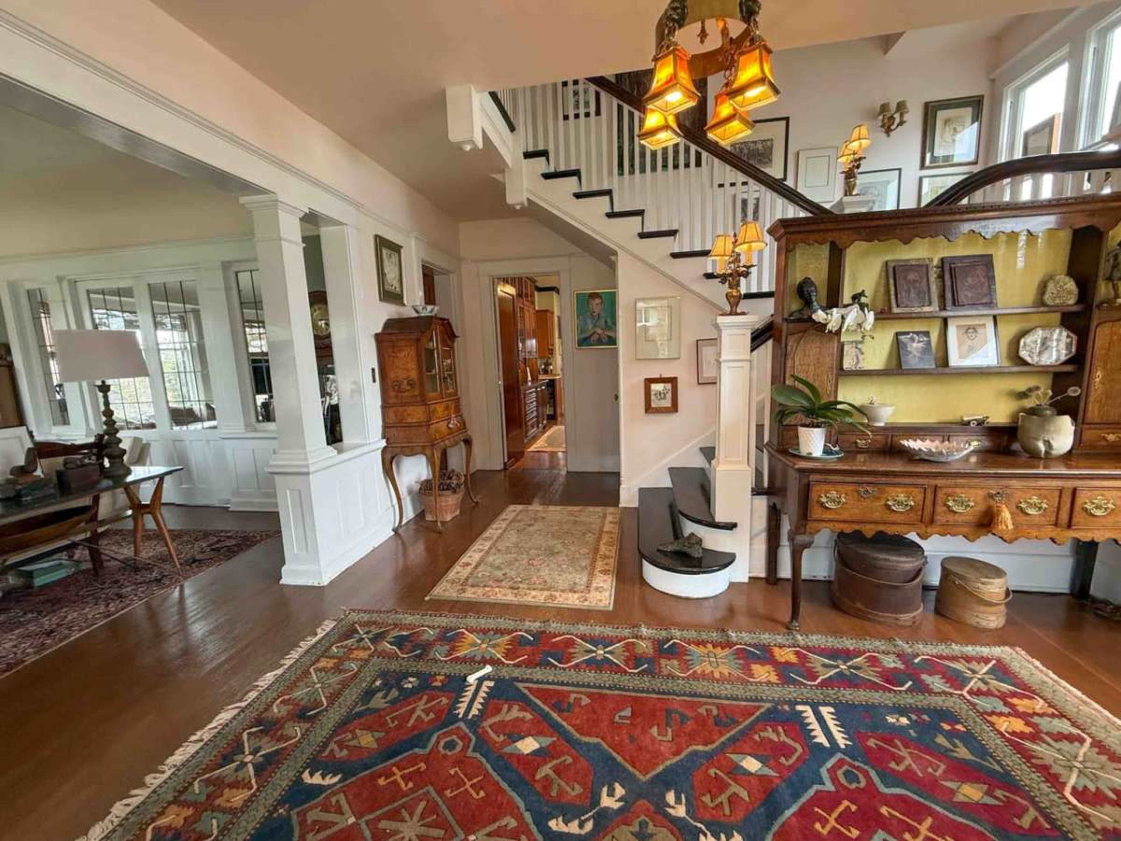The image shows a spacious hallway with wooden floors, a staircase, and various pieces of furniture displayed against the walls.