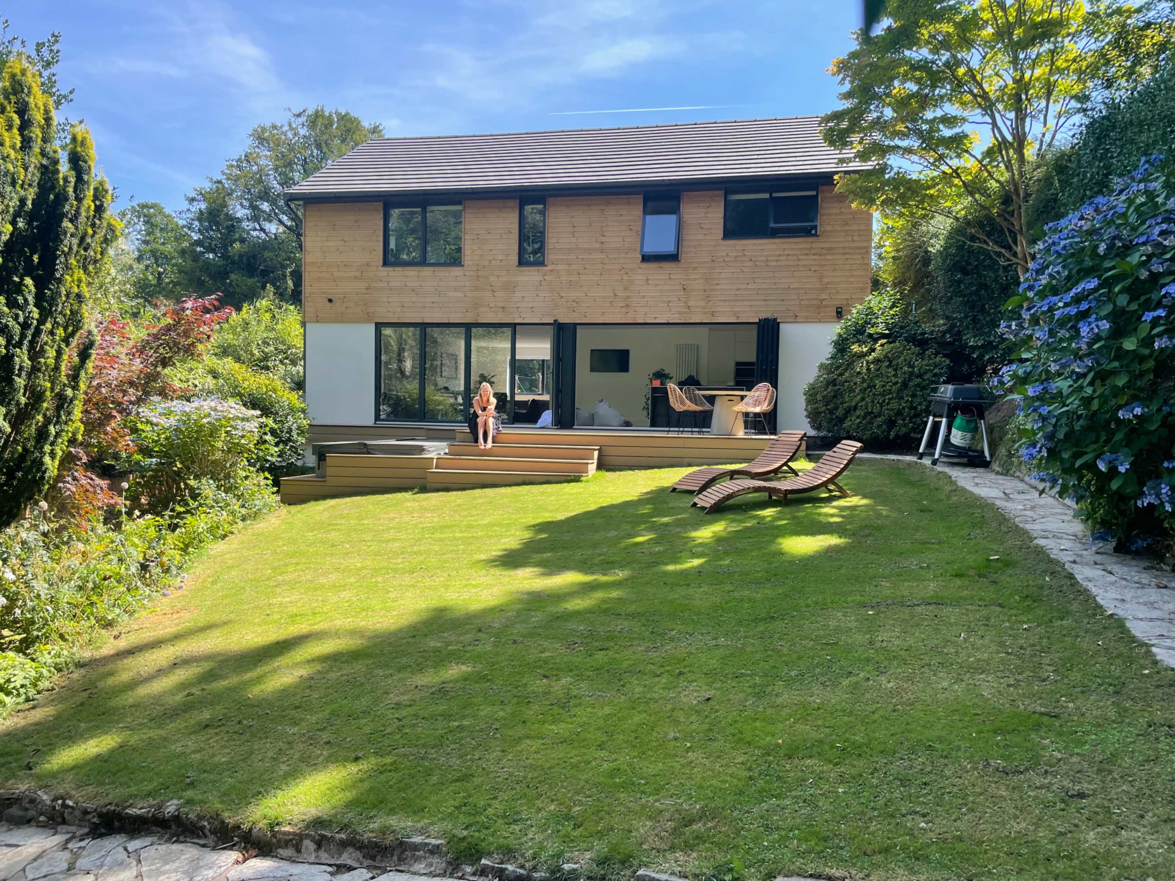 A modern, two-story wooden house overlooks a well-maintained lawn and garden with lounge chairs in the foreground.