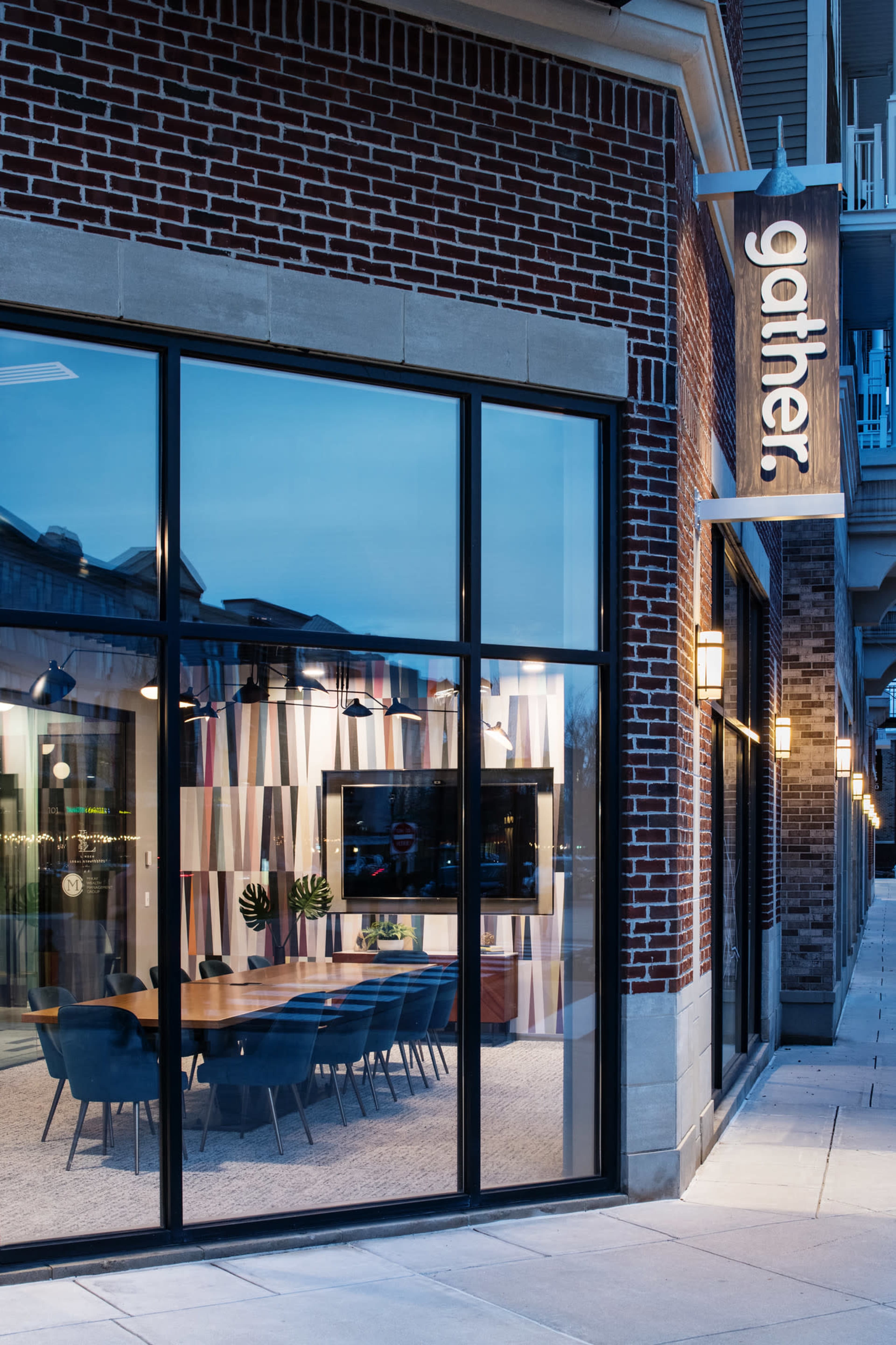 The modern exterior of a building with large glass windows revealing a stylish interior meeting space, illuminated at dusk, marked by a sign that reads "gather."