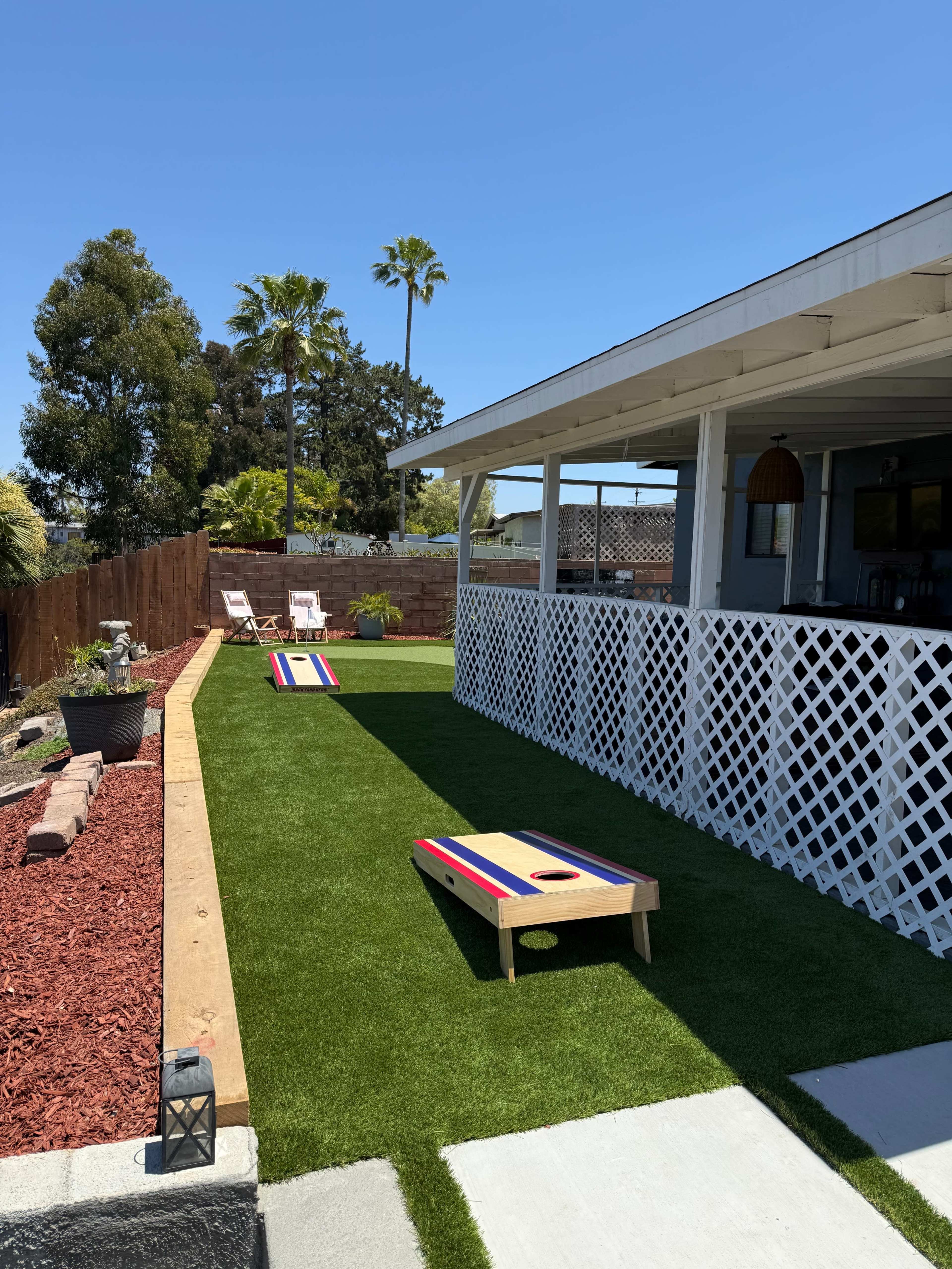 The image shows a backyard with synthetic grass, two cornhole boards set up on the lawn, and white patio chairs beside a white fence.