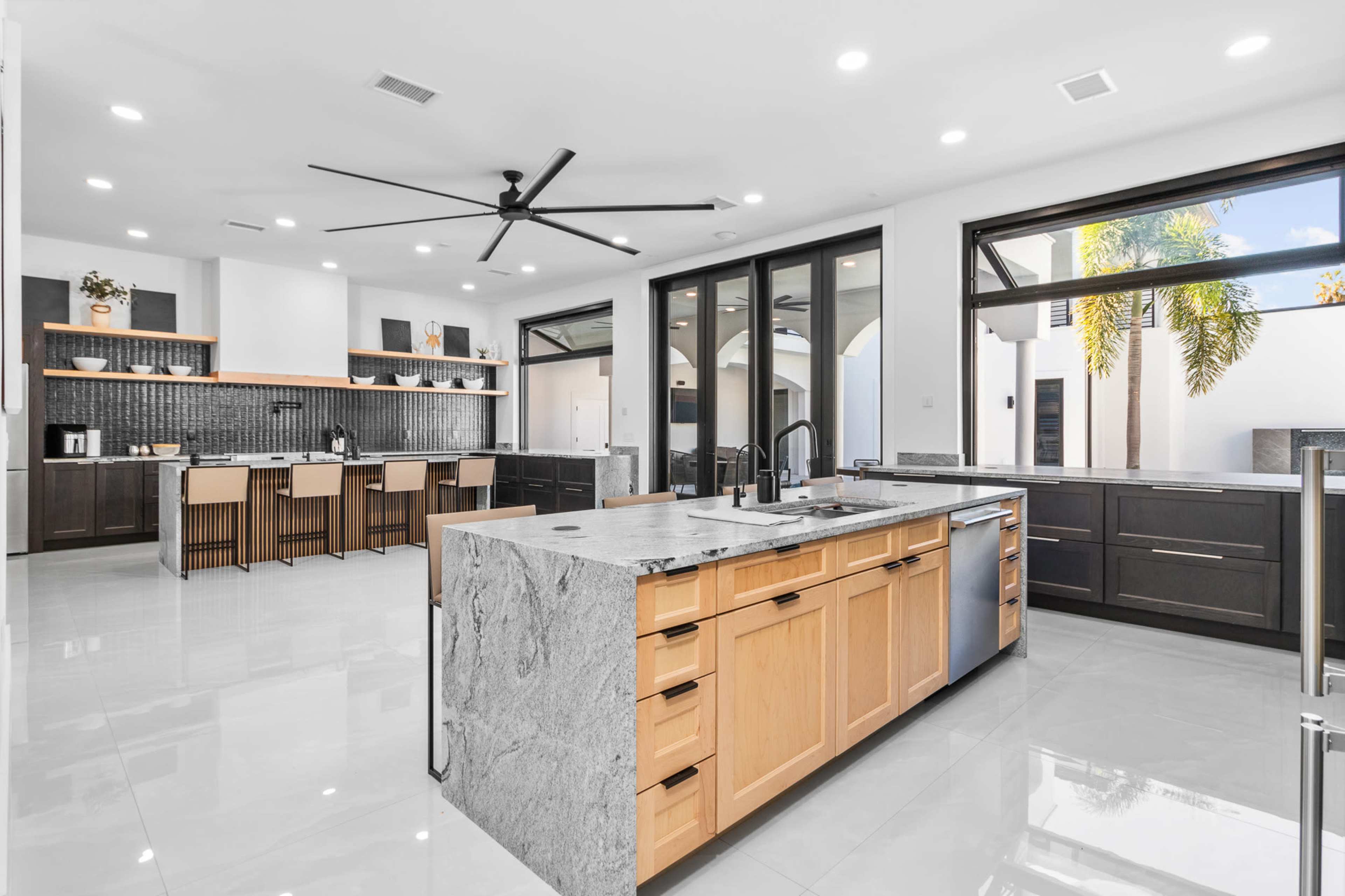 The image shows a modern kitchen with an island featuring a gray marble countertop, dark cabinetry, and large windows allowing natural light to enter the space.