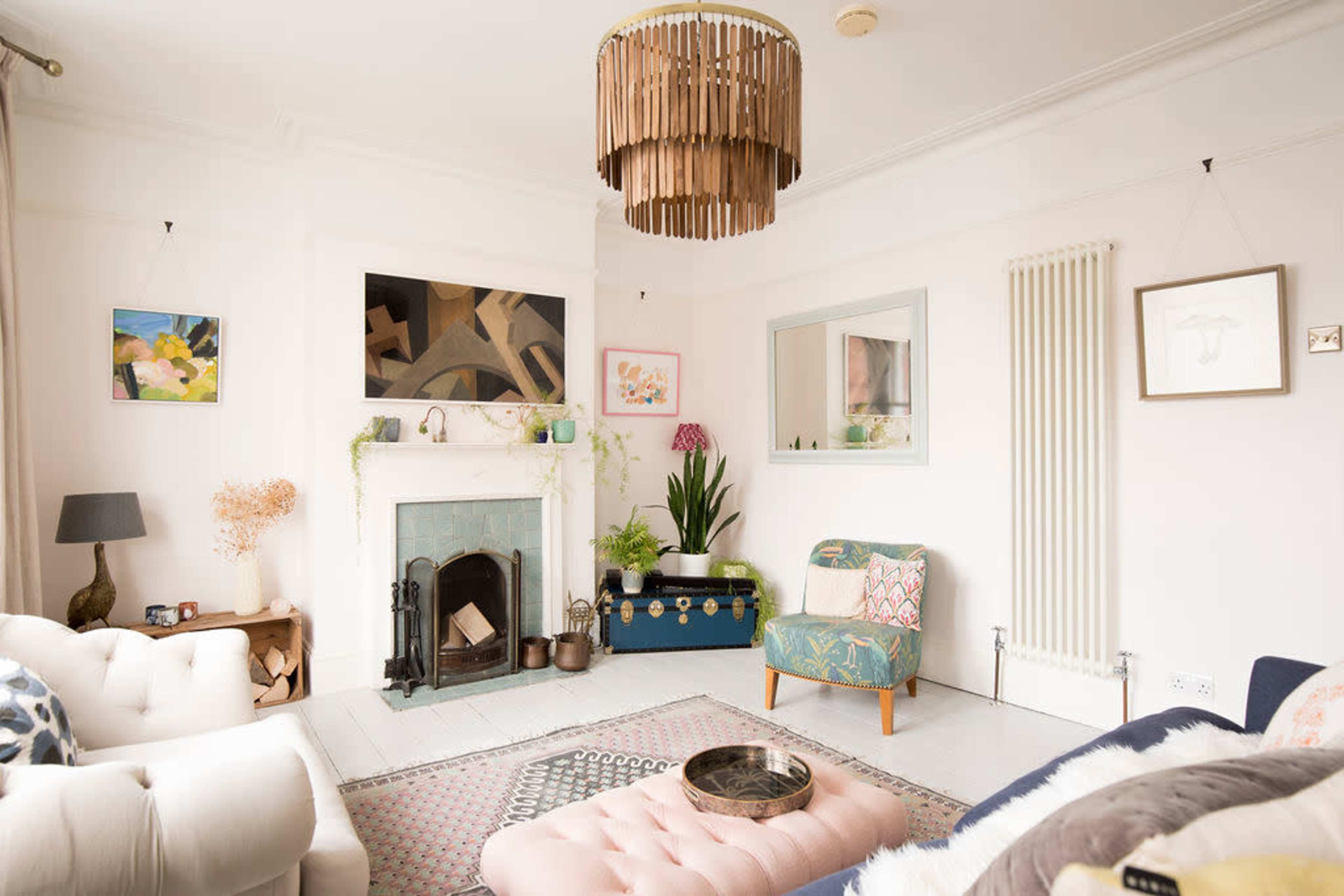 The image shows a bright living room featuring a pastel color scheme, a fireplace with decorative elements, and various pieces of furniture including a tufted ottoman and an accent chair.