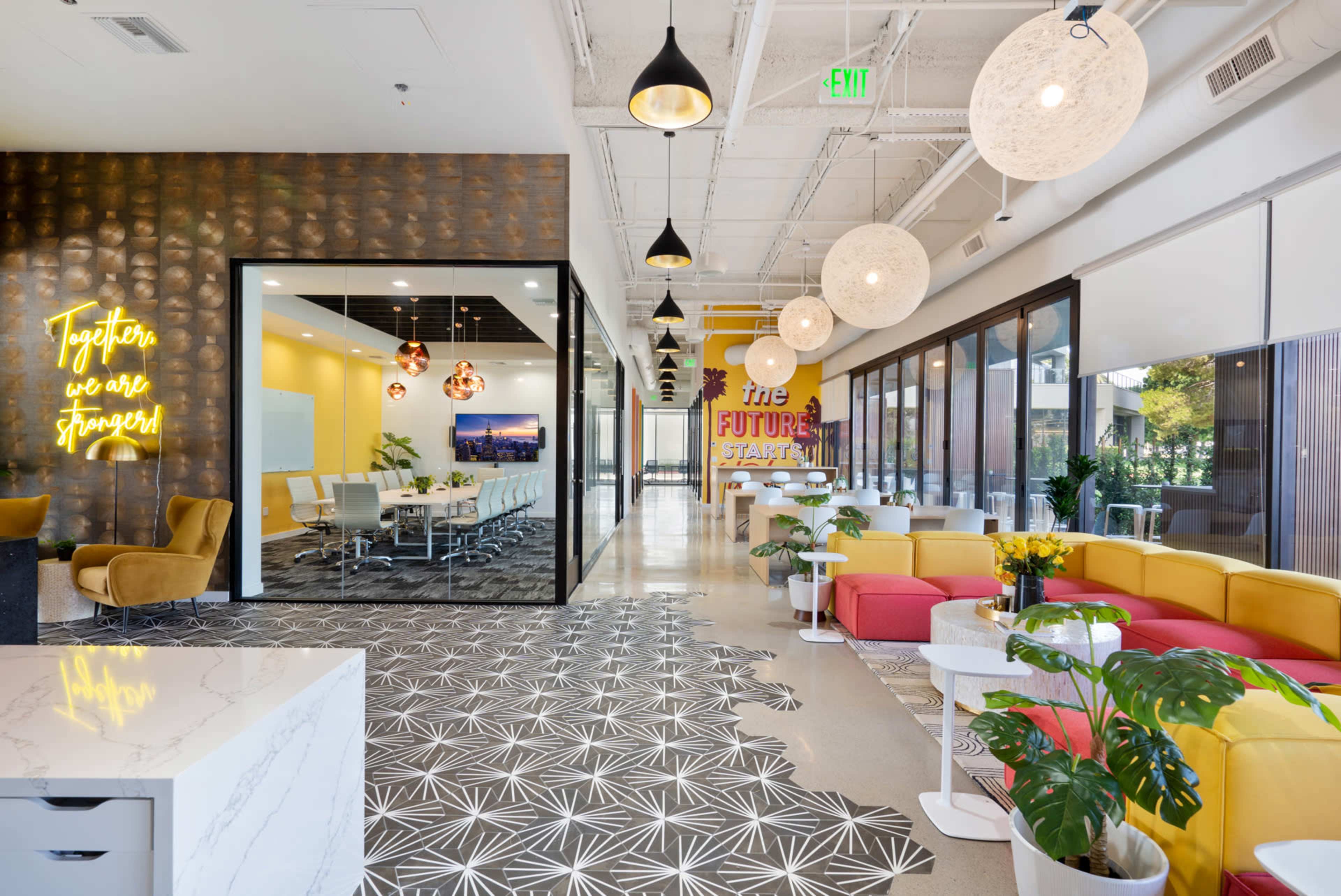 The image shows a modern, open office space featuring vibrant yellow and pink seating areas, a conference room with a glass wall, and decorative lighting.