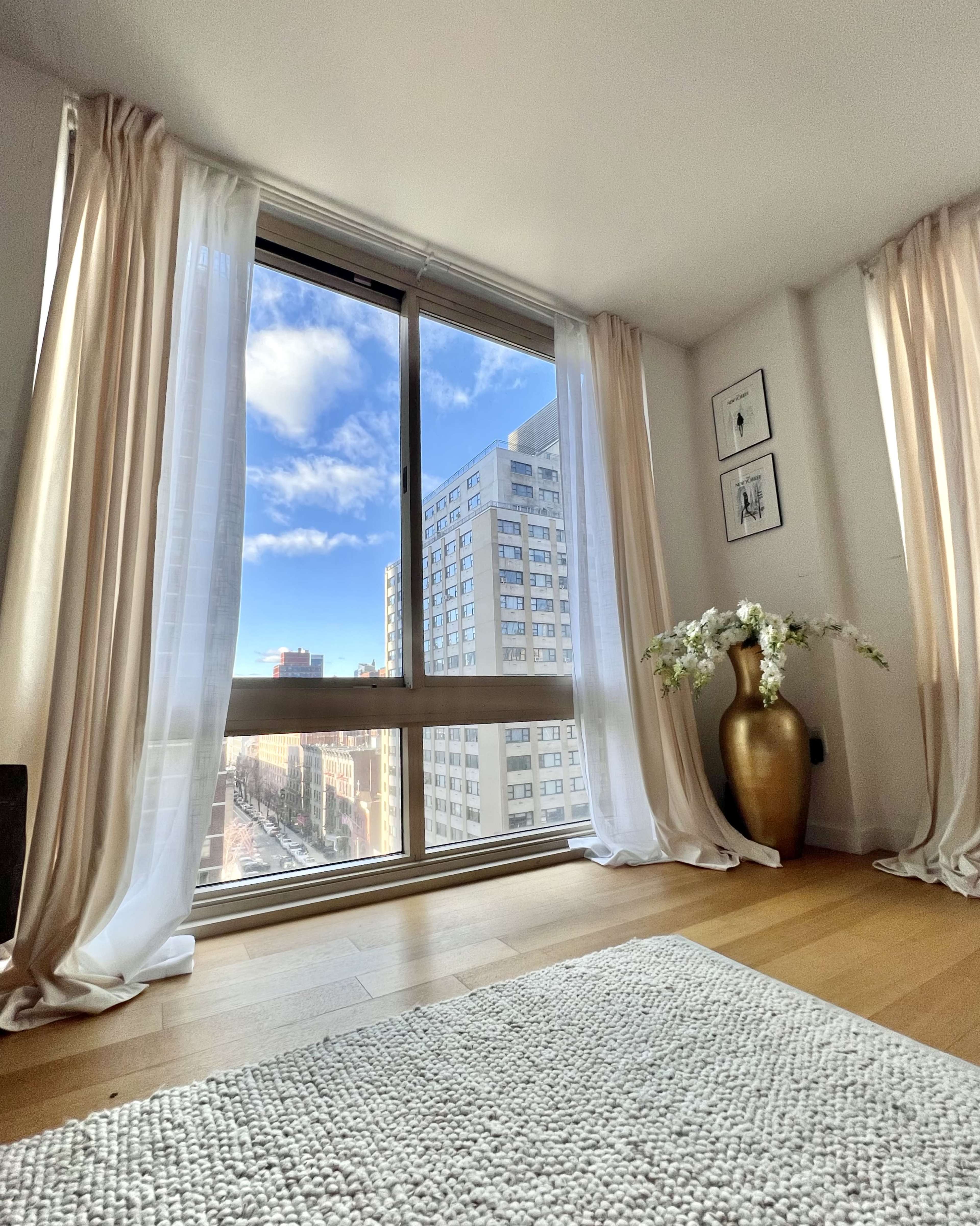 A bright room with large windows overlooks a city street, featuring sheer curtains and a decorative vase.