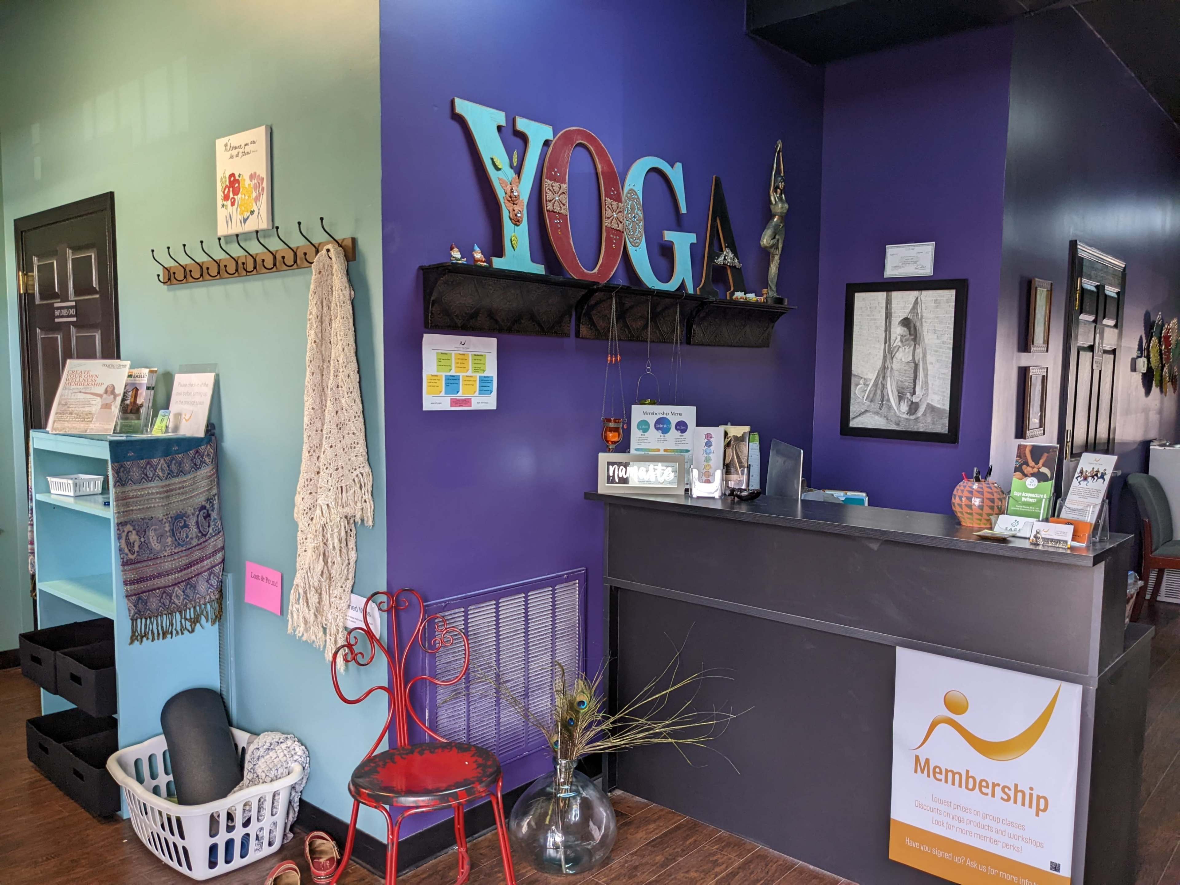 A brightly colored yoga studio reception area featuring a large "YOGA" sign, a black counter with membership information, and various decorative elements on the walls.