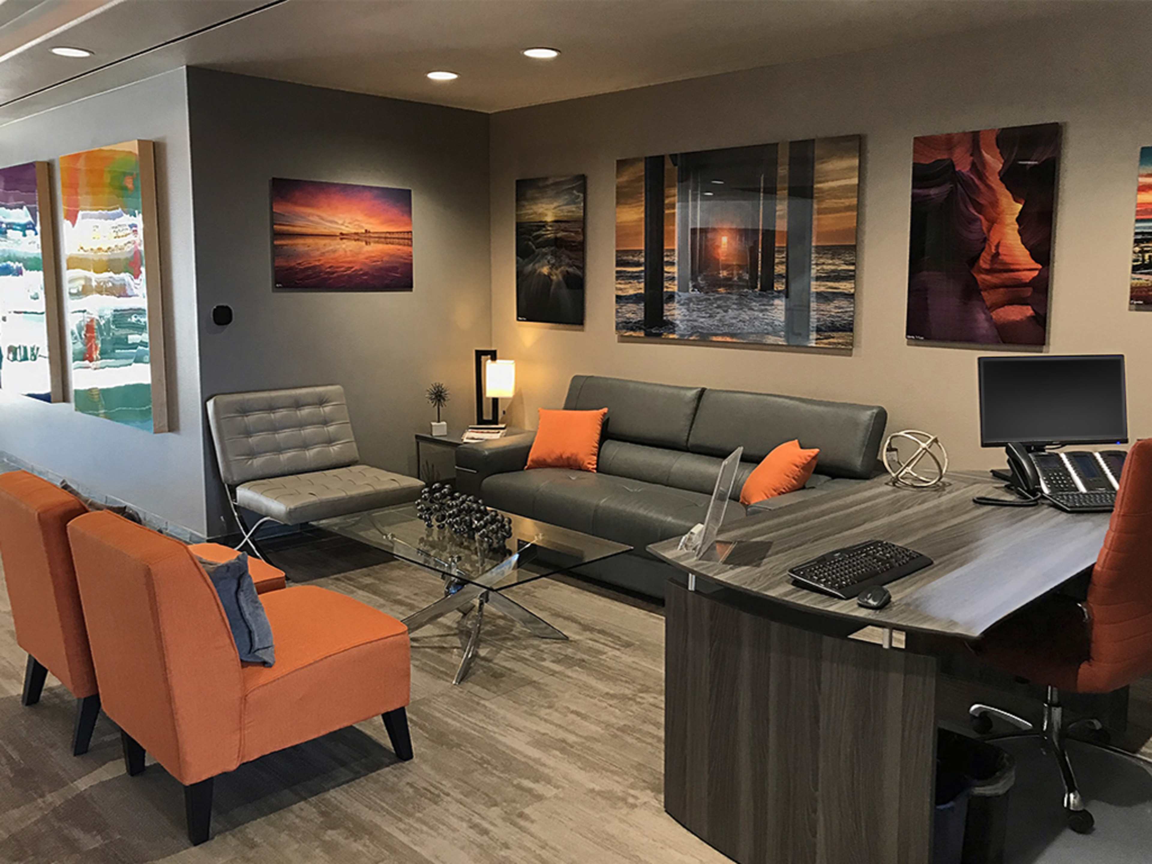 A modern office reception area with gray furniture, orange accent chairs, and framed artwork on the walls.