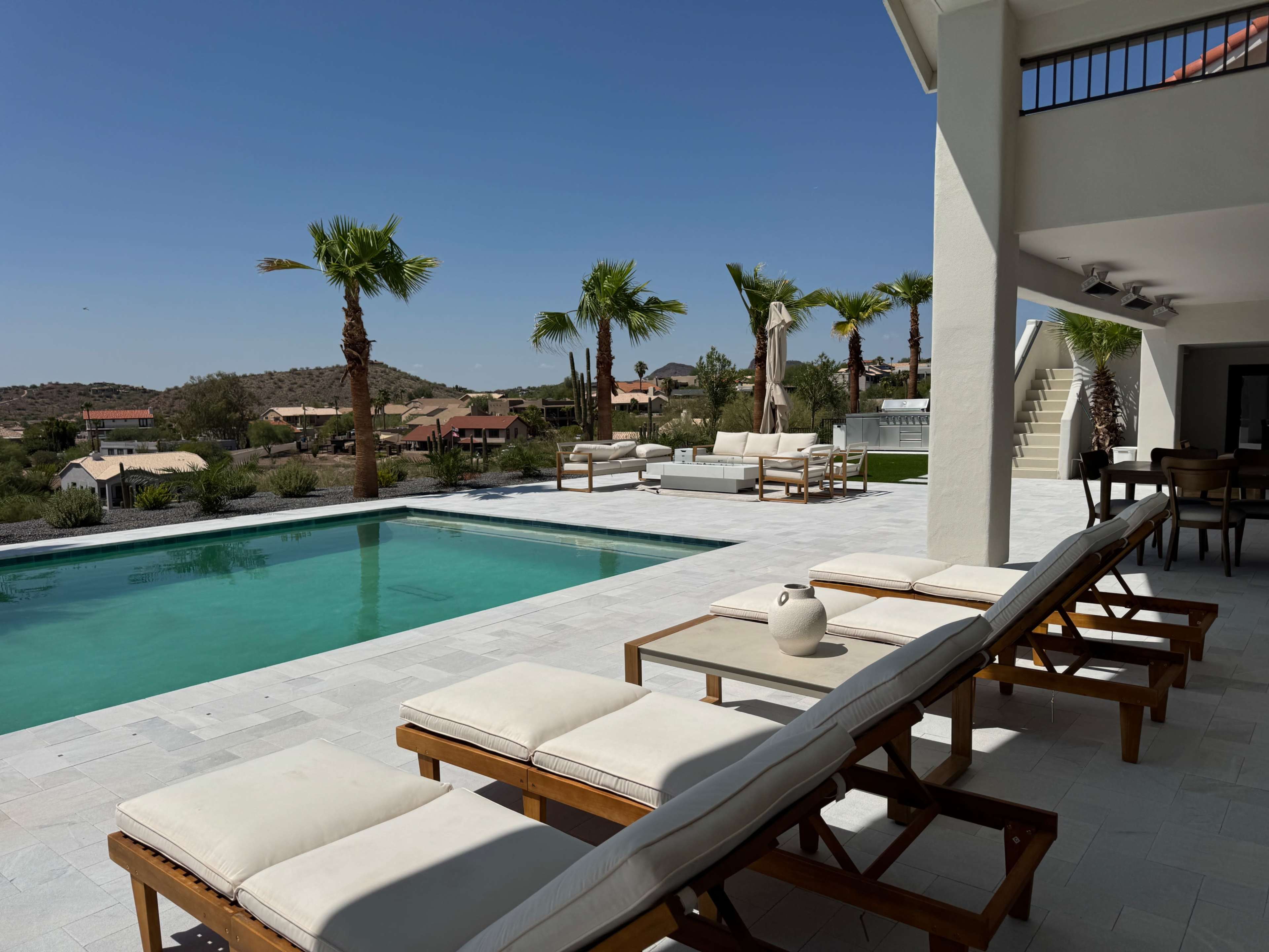 A modern villa with a swimming pool and lounge chairs overlooks a desert landscape dotted with palm trees.