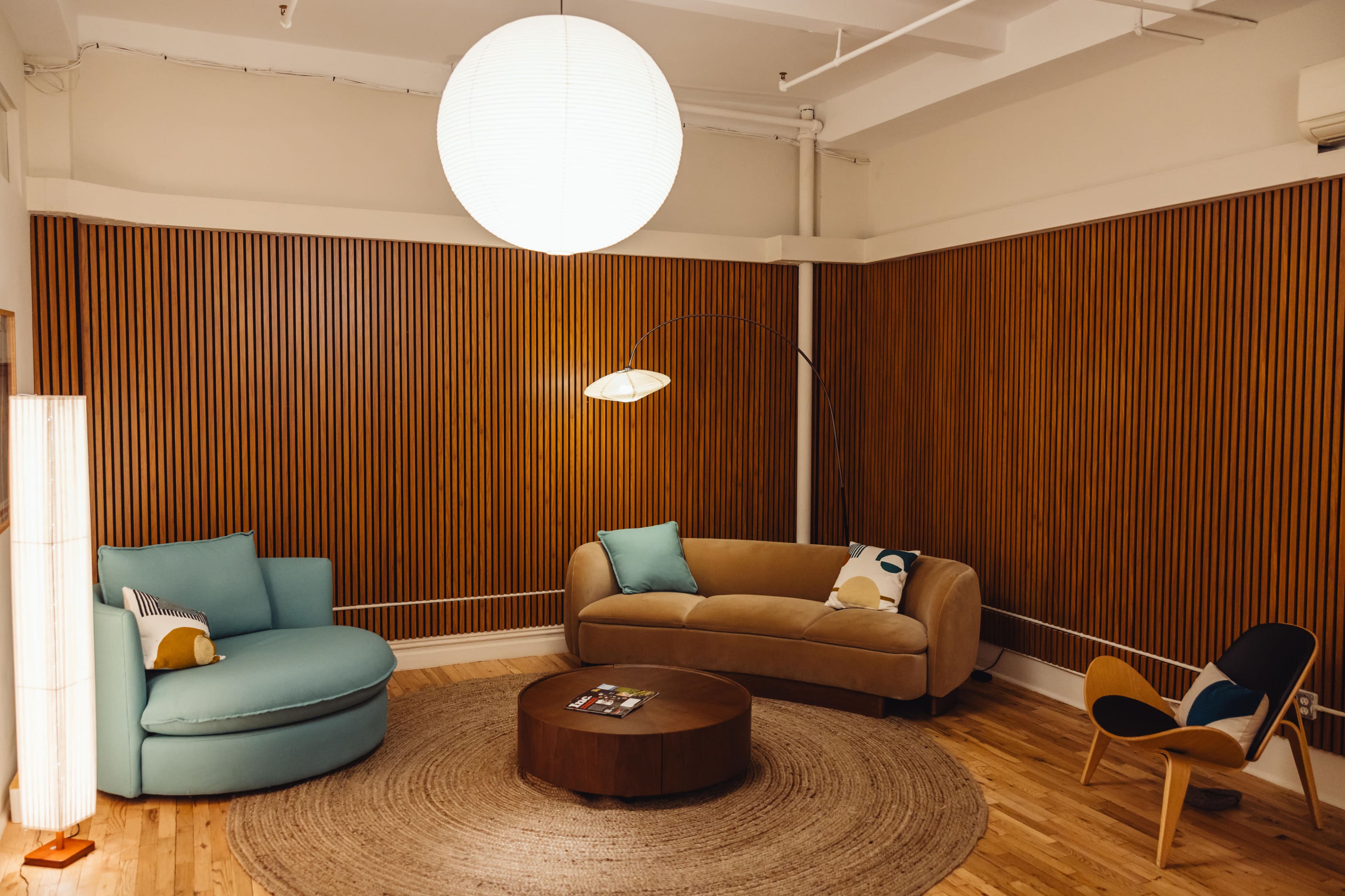 The image features a cozy seating area with a blue lounge chair, a beige curved sofa, a circular wooden coffee table, and a large pendant light, all set against a backdrop of wooden paneling.