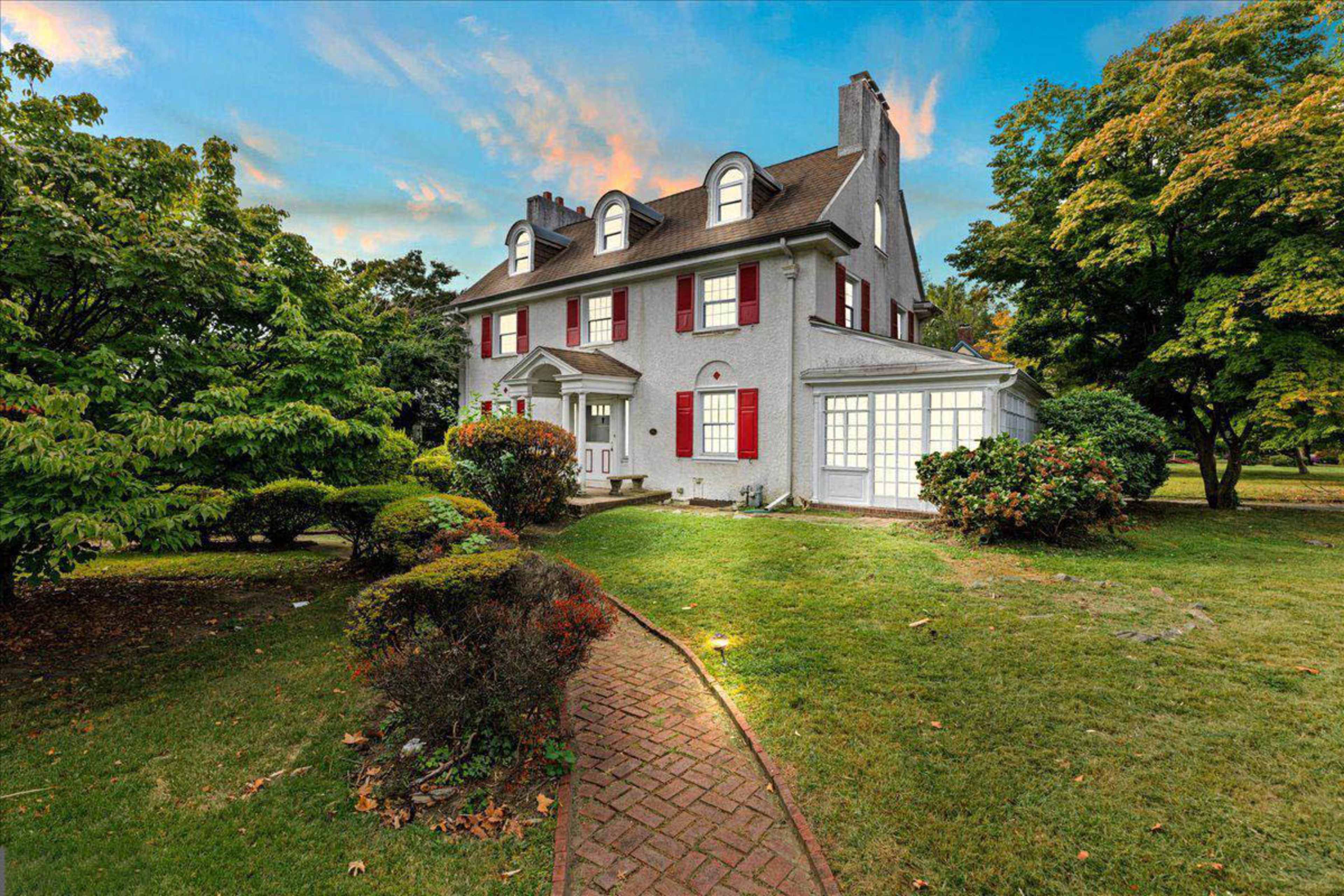 The image shows a two-story house with a gray exterior, red shutters, and a landscaped yard featuring shrubs and trees.