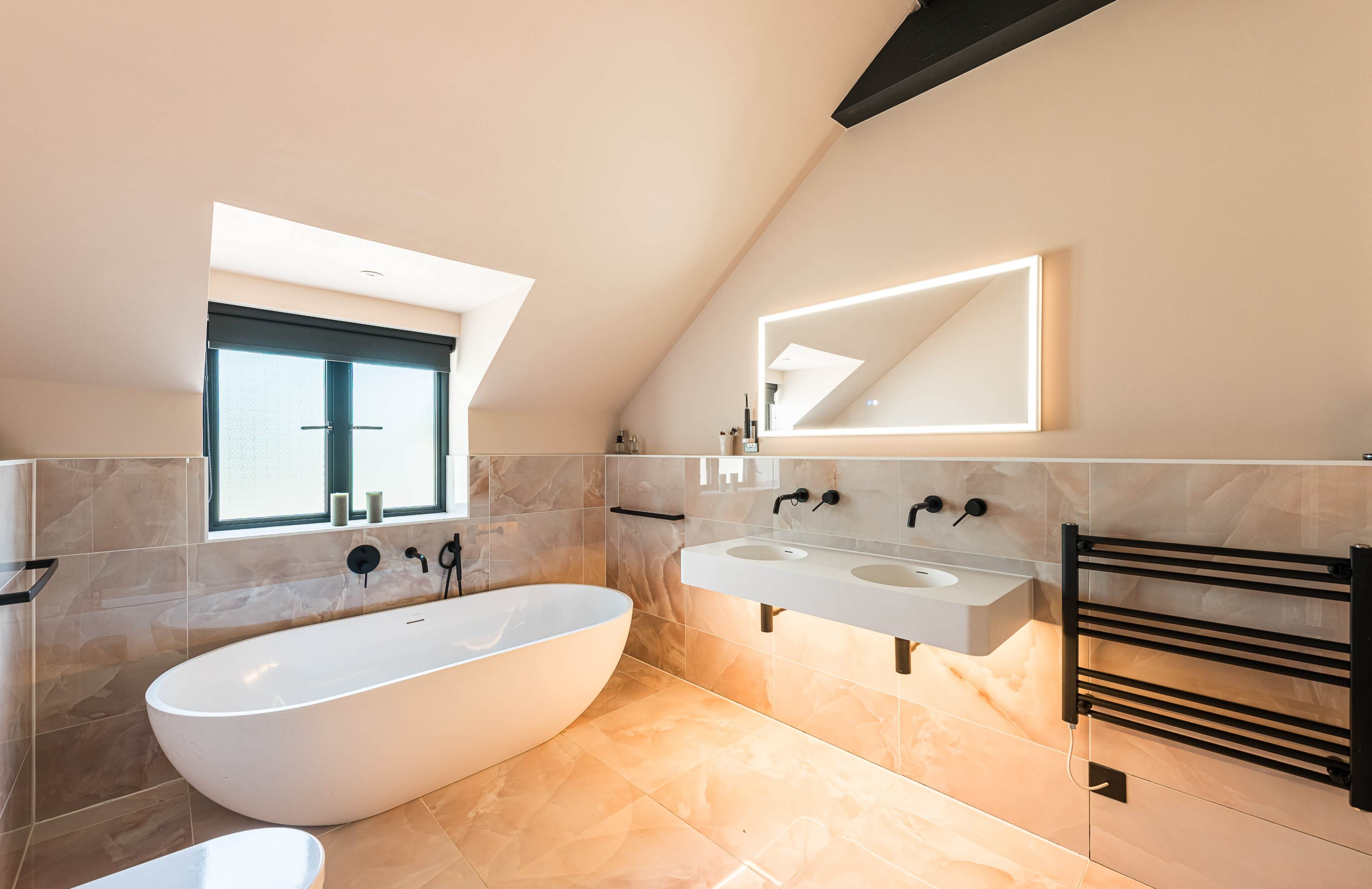 The image shows a modern bathroom with a freestanding white bathtub, dual sinks on a counter, and a large mirror illuminated by backlighting.