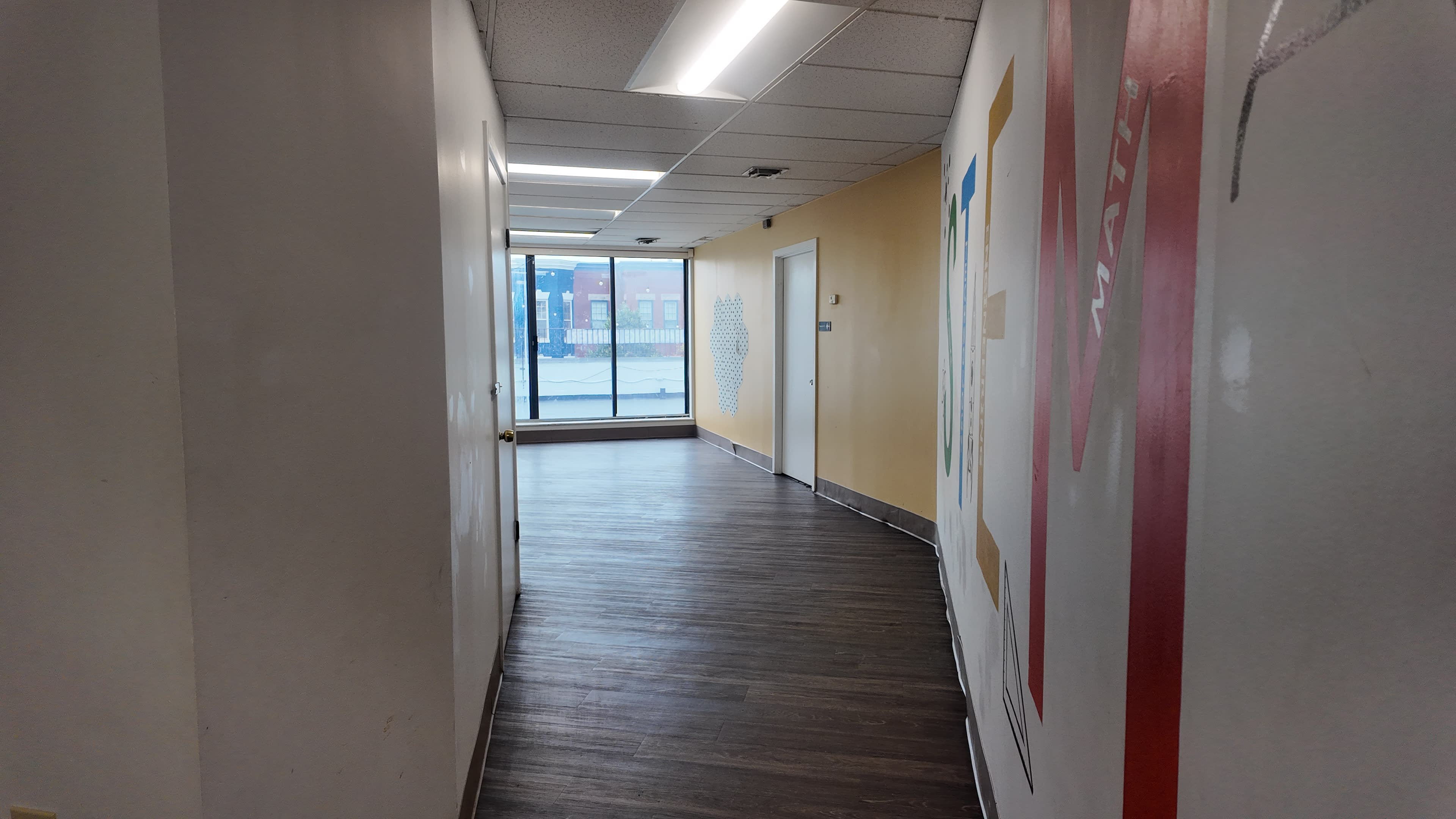 The image shows a hallway with a wooden floor, light-colored walls, and a large window at the end that reveals an outdoor view.