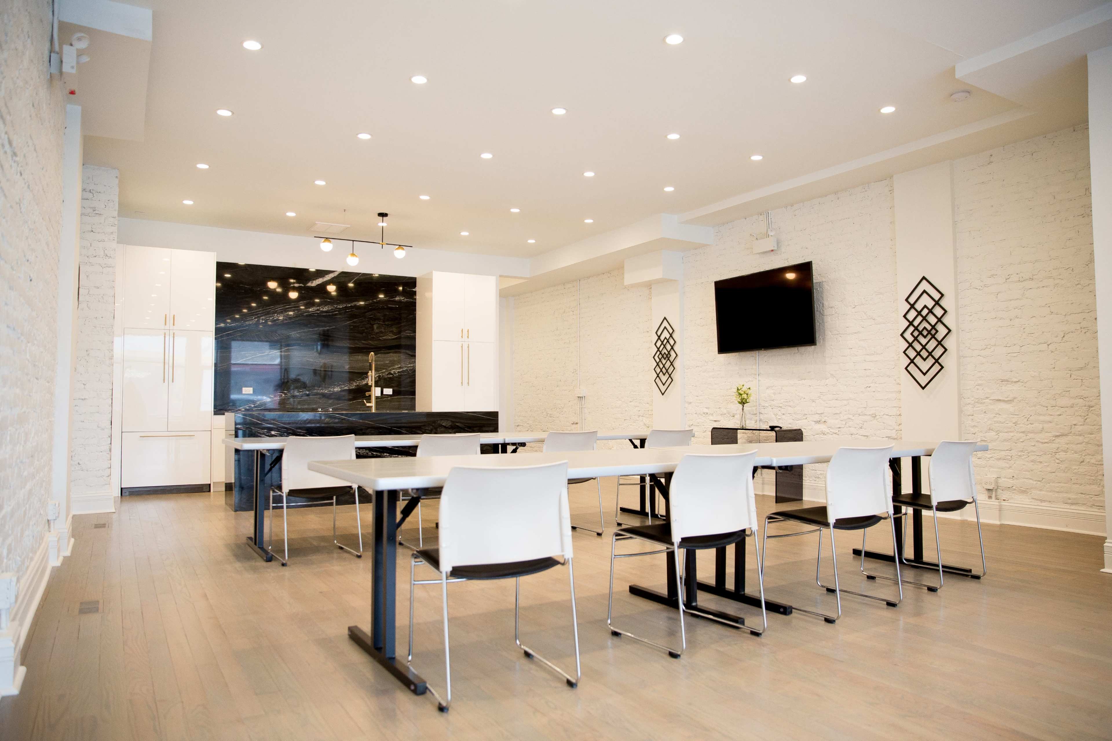 A modern conference room features a long table with white chairs, a wall-mounted TV, and a black kitchen area against a white brick backdrop.