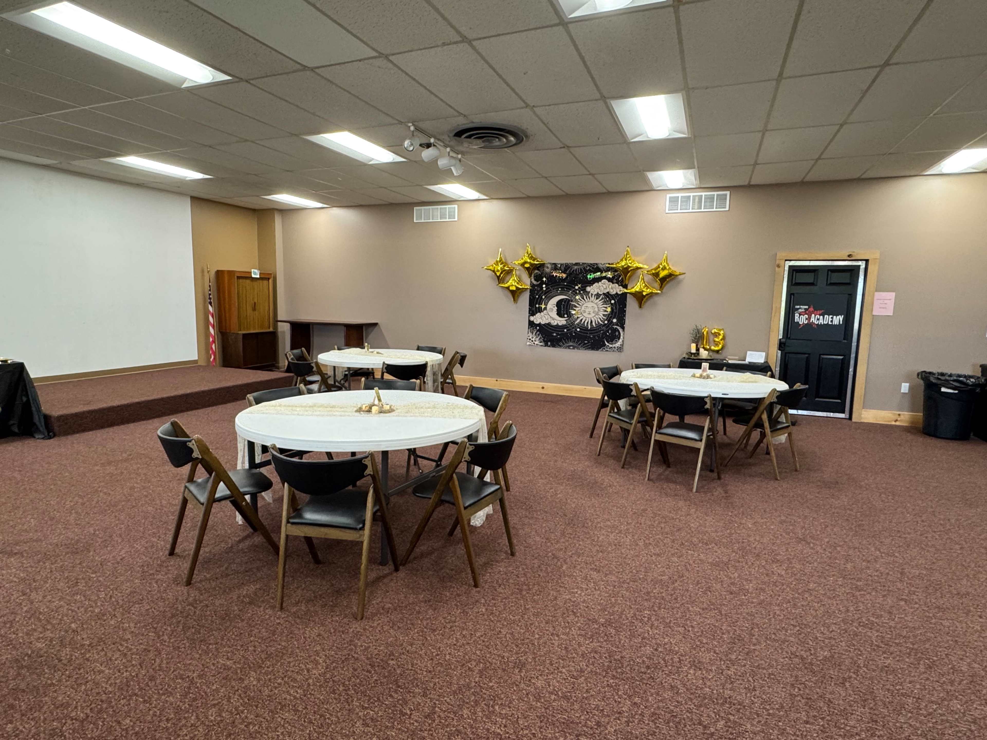 The image shows a spacious room set up with several round tables and chairs, decorated with gold star balloons on the wall.