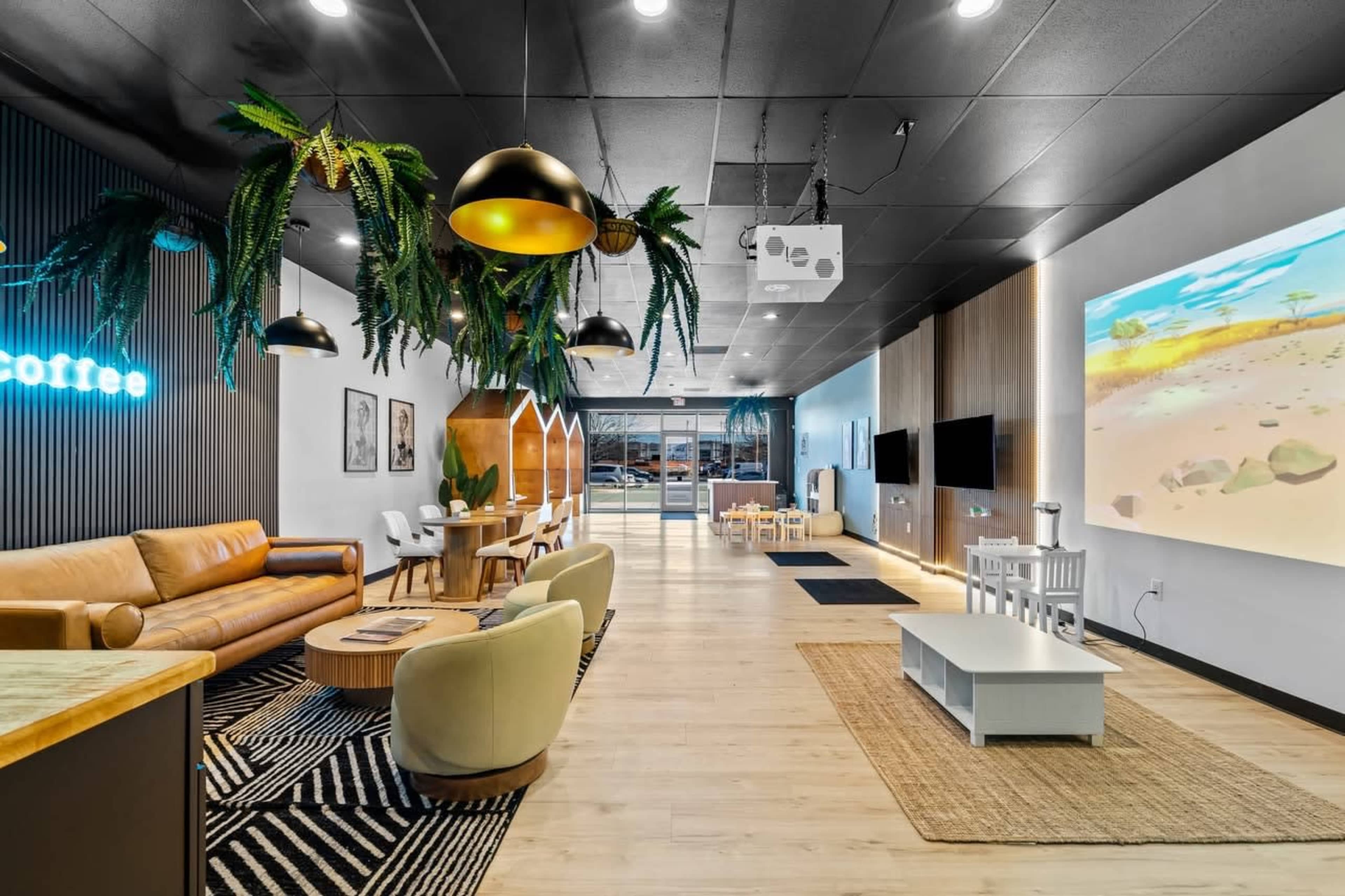 The image shows a modern coffee shop interior with a mix of seating, decorative plants, and a large screen displaying an animated scene.