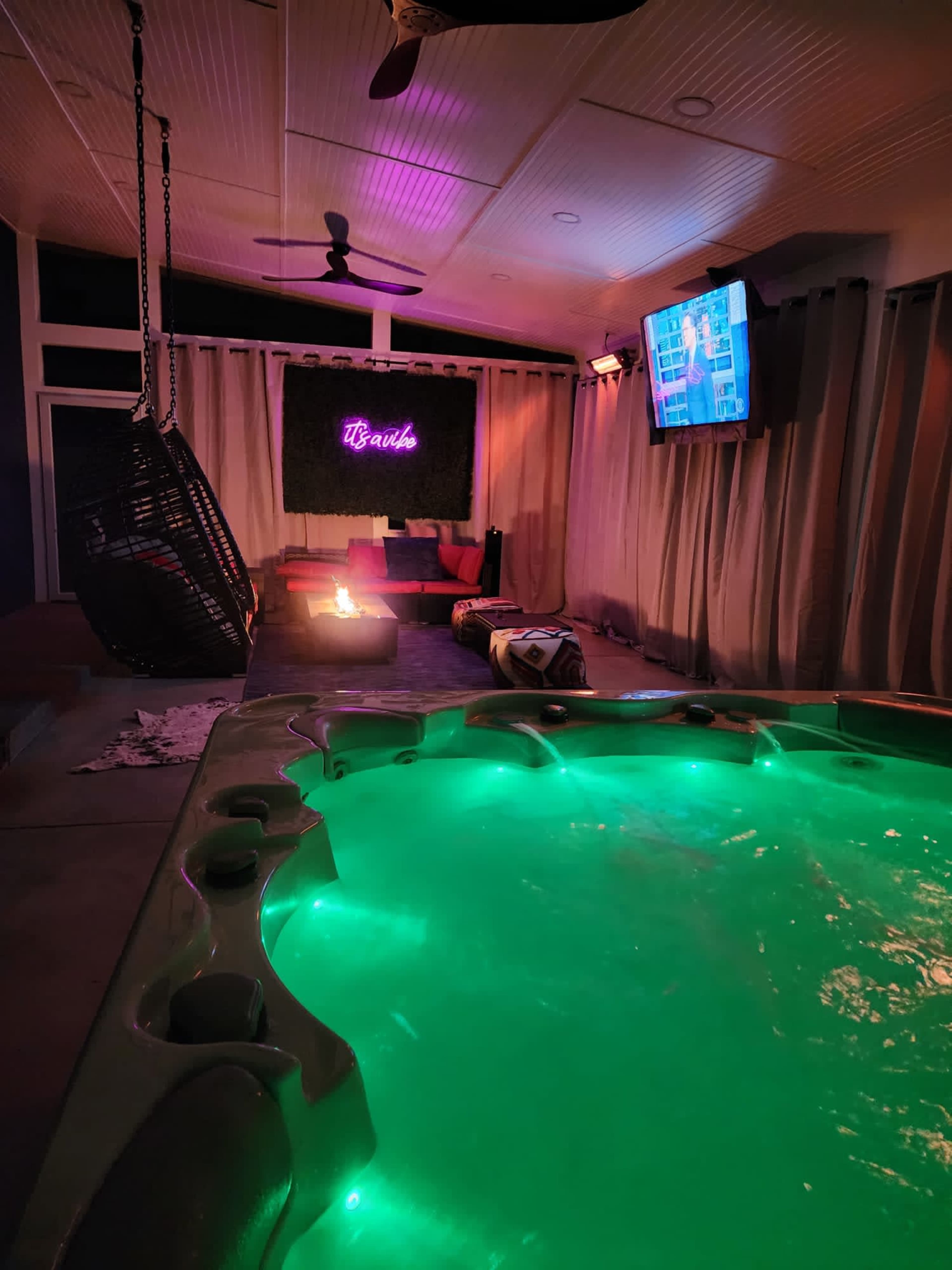 A lit hot tub with green water is set in a cozy indoor space featuring a hanging chair, a television, and a fire pit surrounded by dim lighting.