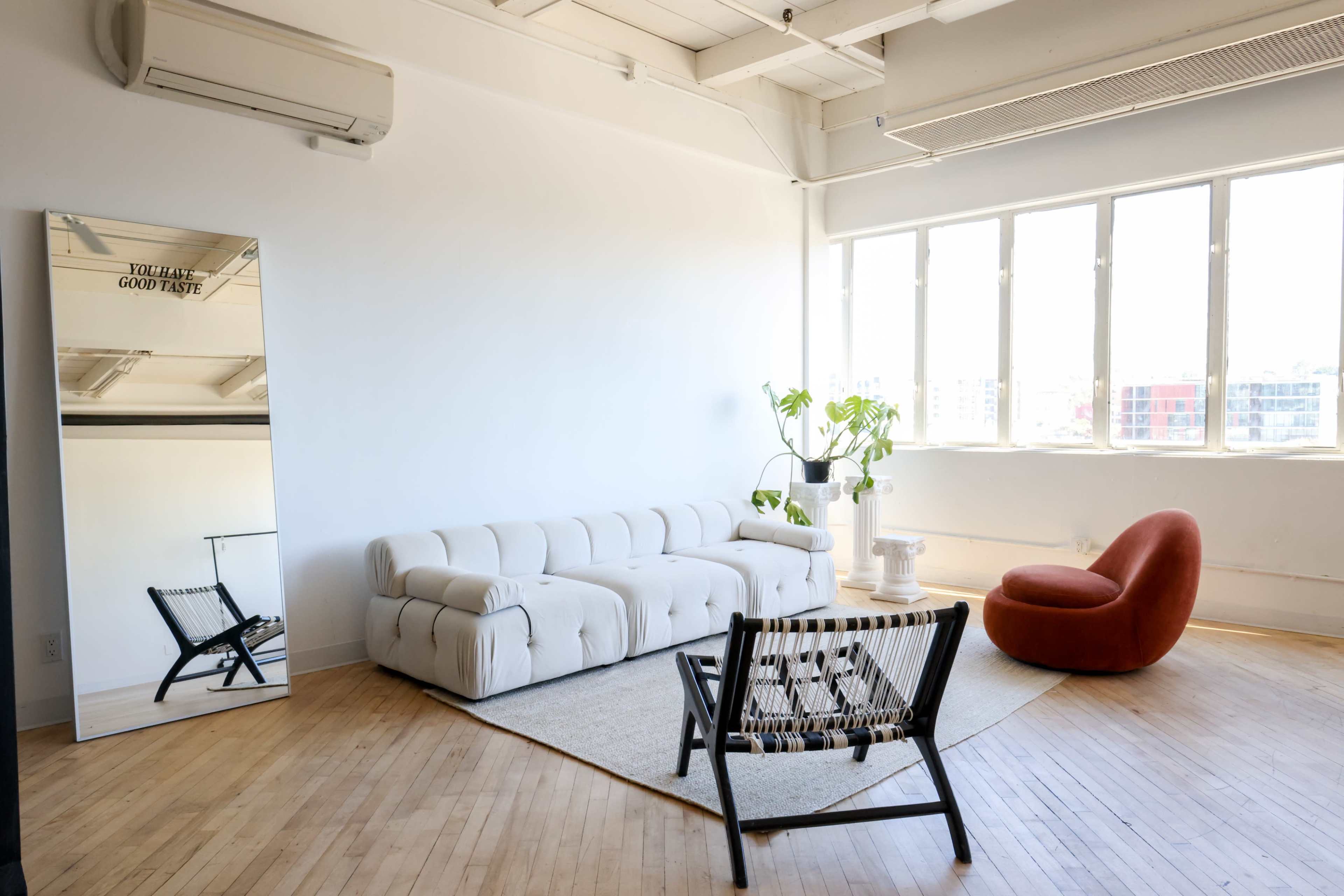 A spacious room with hardwood floors, featuring a beige sofa, a red accent chair, a black woven chair, a white side table, and a large mirror reflecting the sunlight from the windows.