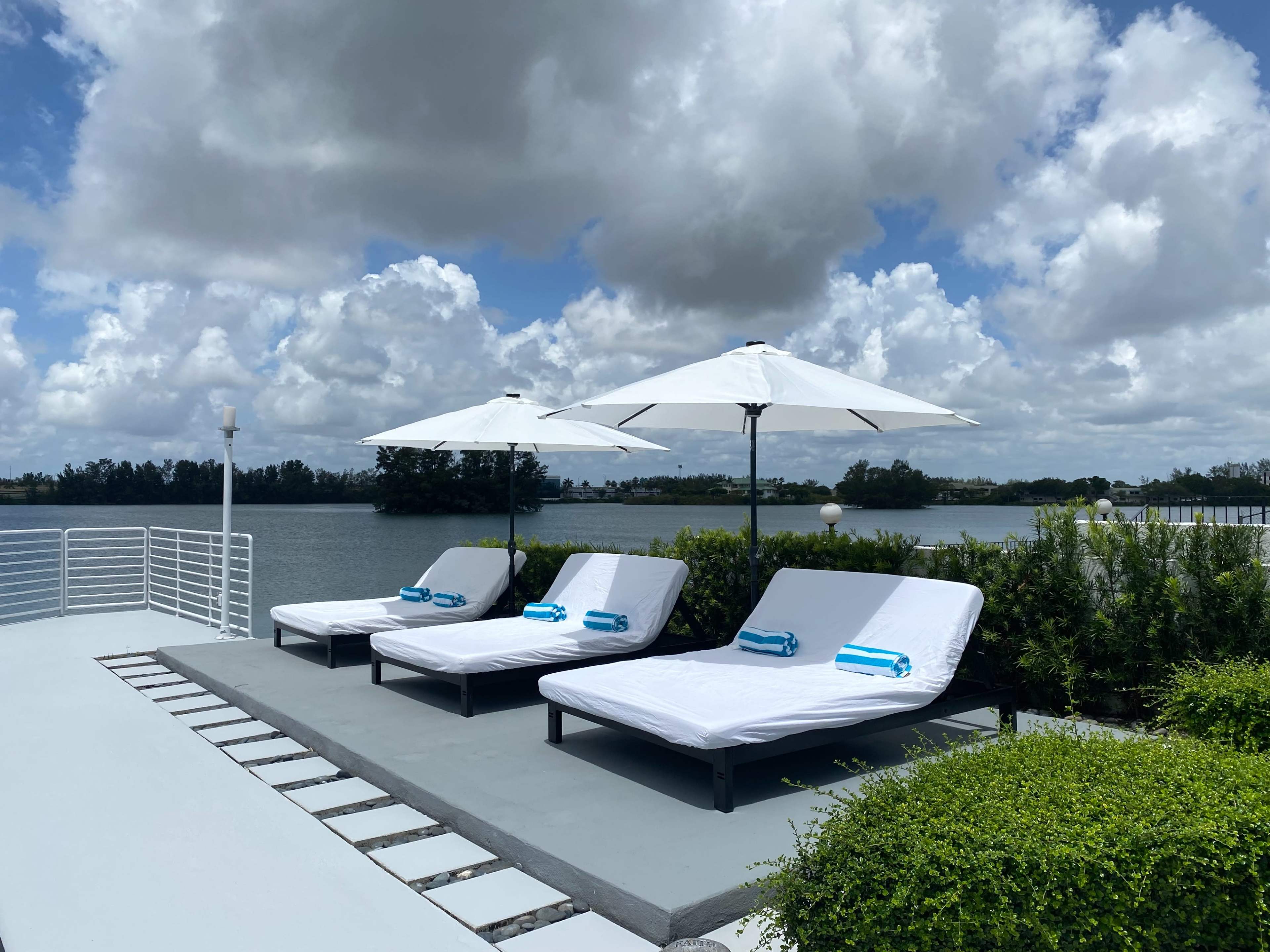 Three lounge chairs with blue towels sit under white umbrellas by a river with greenery in the background.
