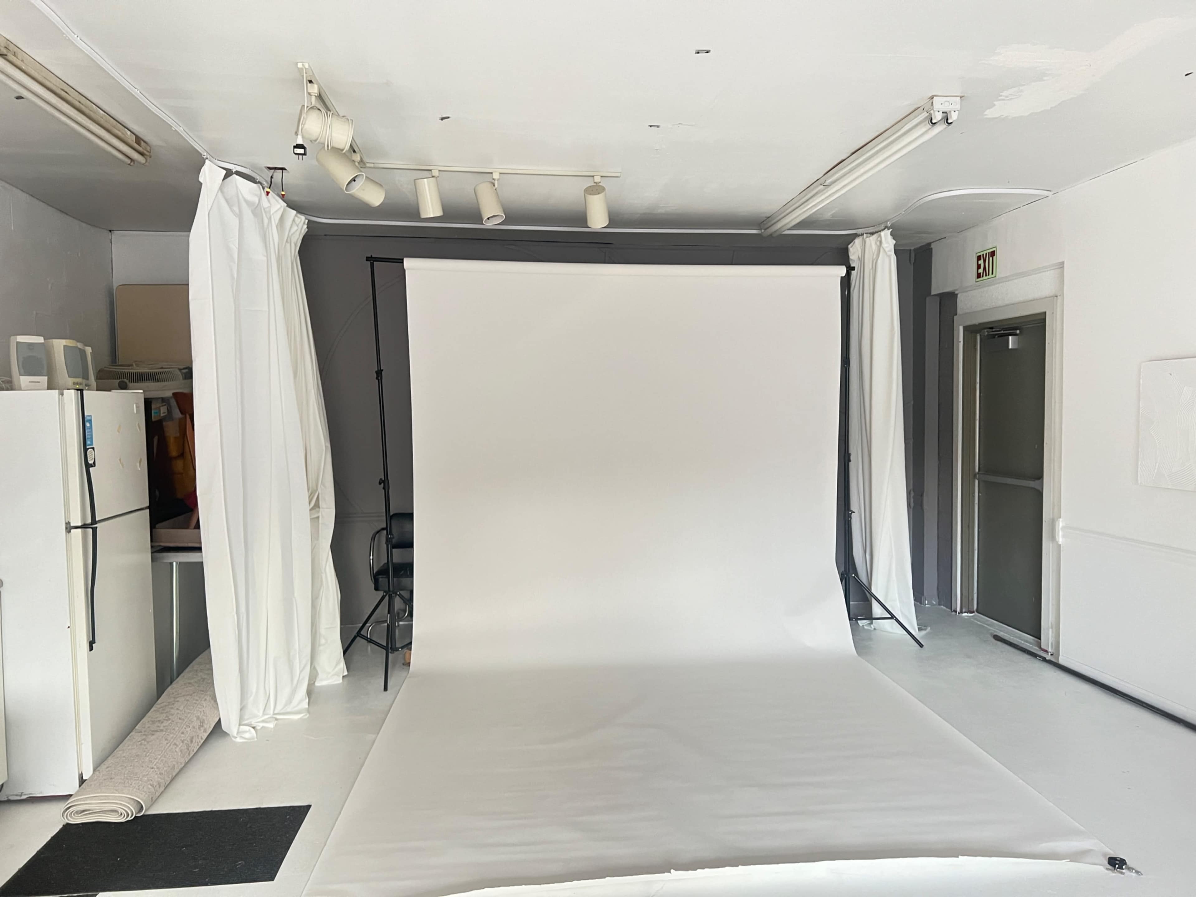 A photography studio with a white backdrop, a small refrigerator, and white curtains on the walls.