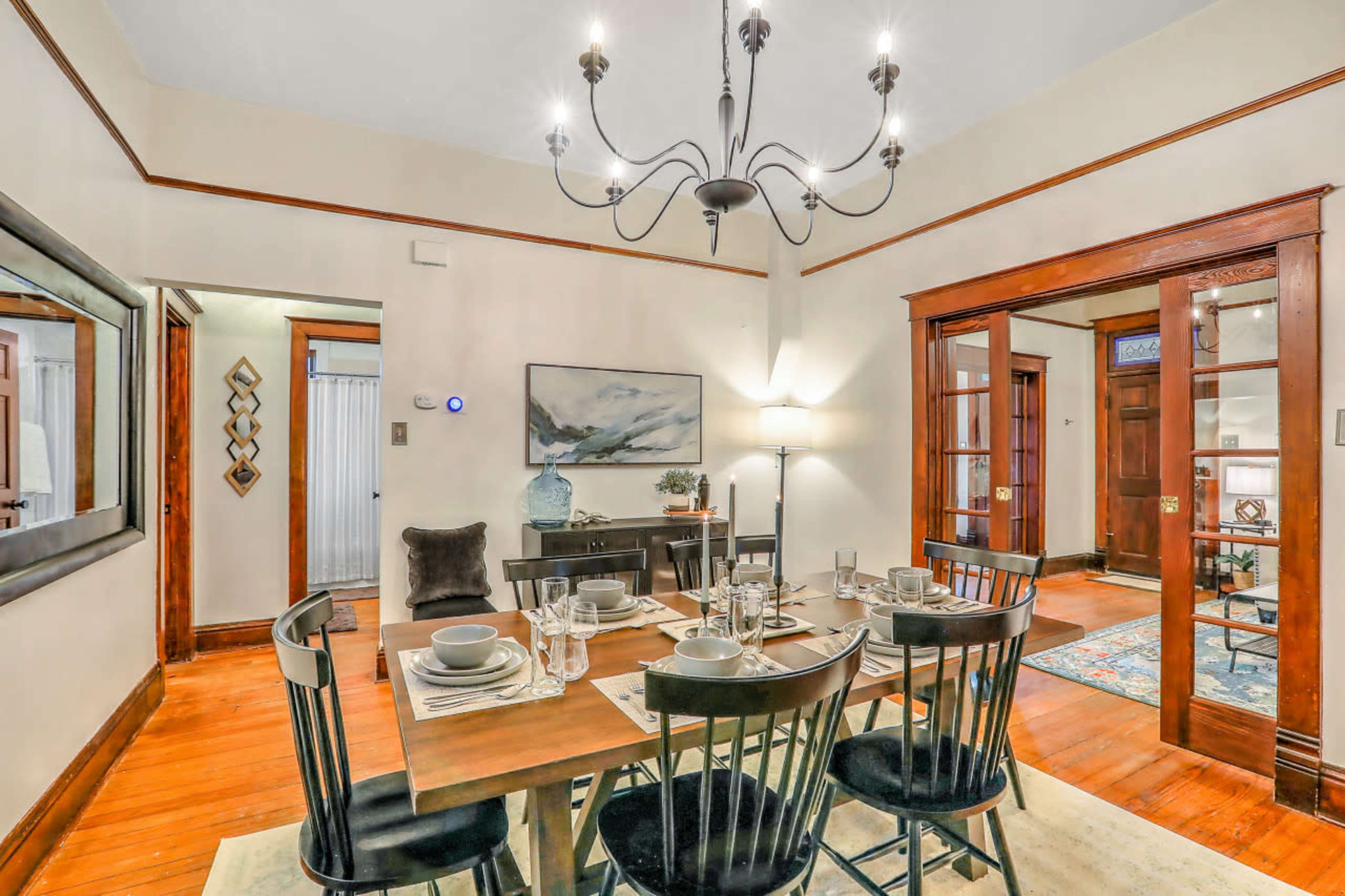 A dining room features a wooden table set for six with plates and glasses, illuminated by a chandelier, and adjacent to a living area through glass-paneled doors.
